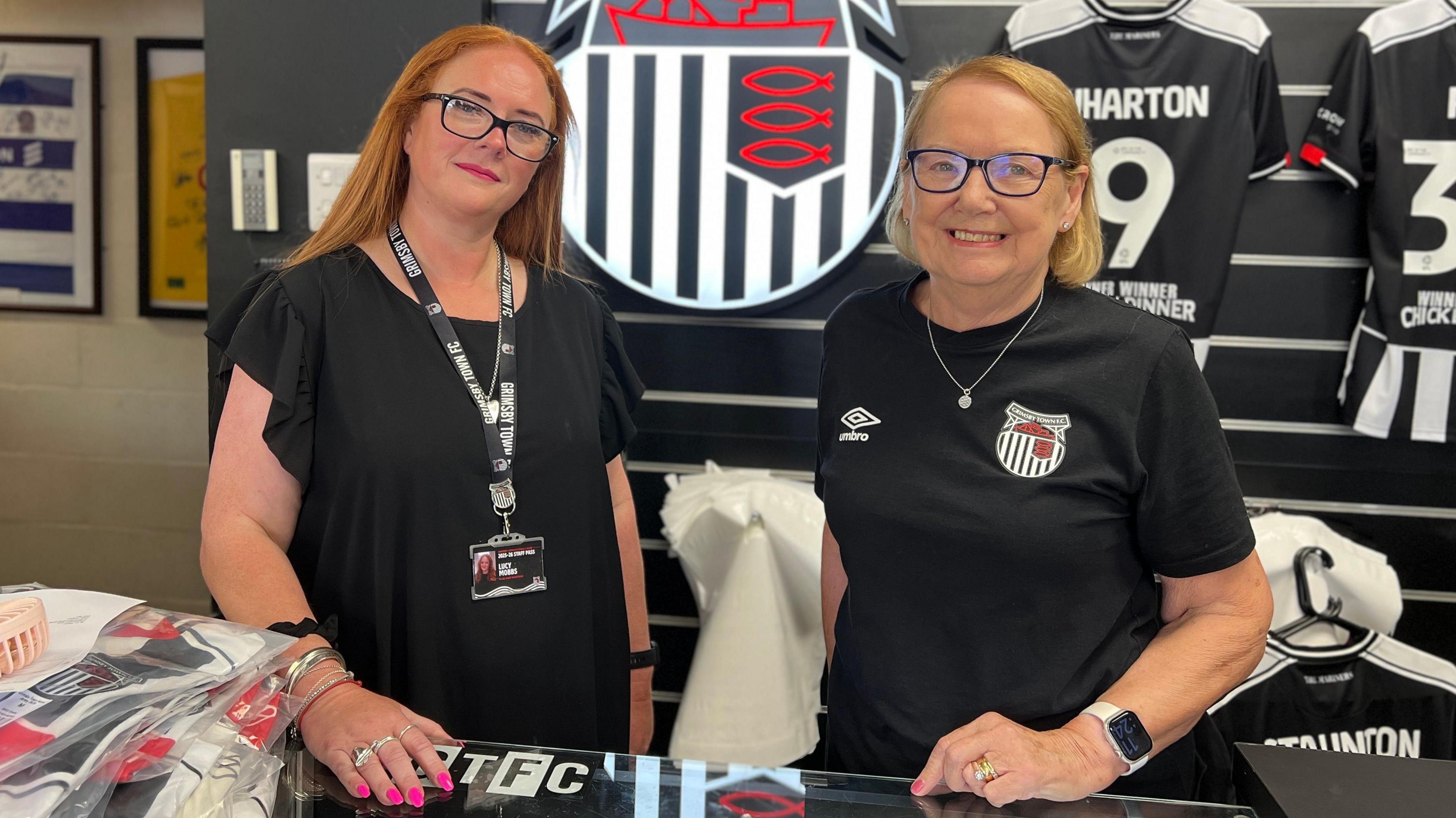 Two women stand inside the Grimsby Town FC merchandise shop. One has ginger hair and wears a black dress, lanyard and glasses; the other has blonde hair and wears a black club-branded T-shirt and glasses. Behind them, a large Grimsby Town crest is displayed on the wall, flanked by a football shirt labelled "Wharton 19". Club merchandise including shirts and scarves is arranged on the counter.