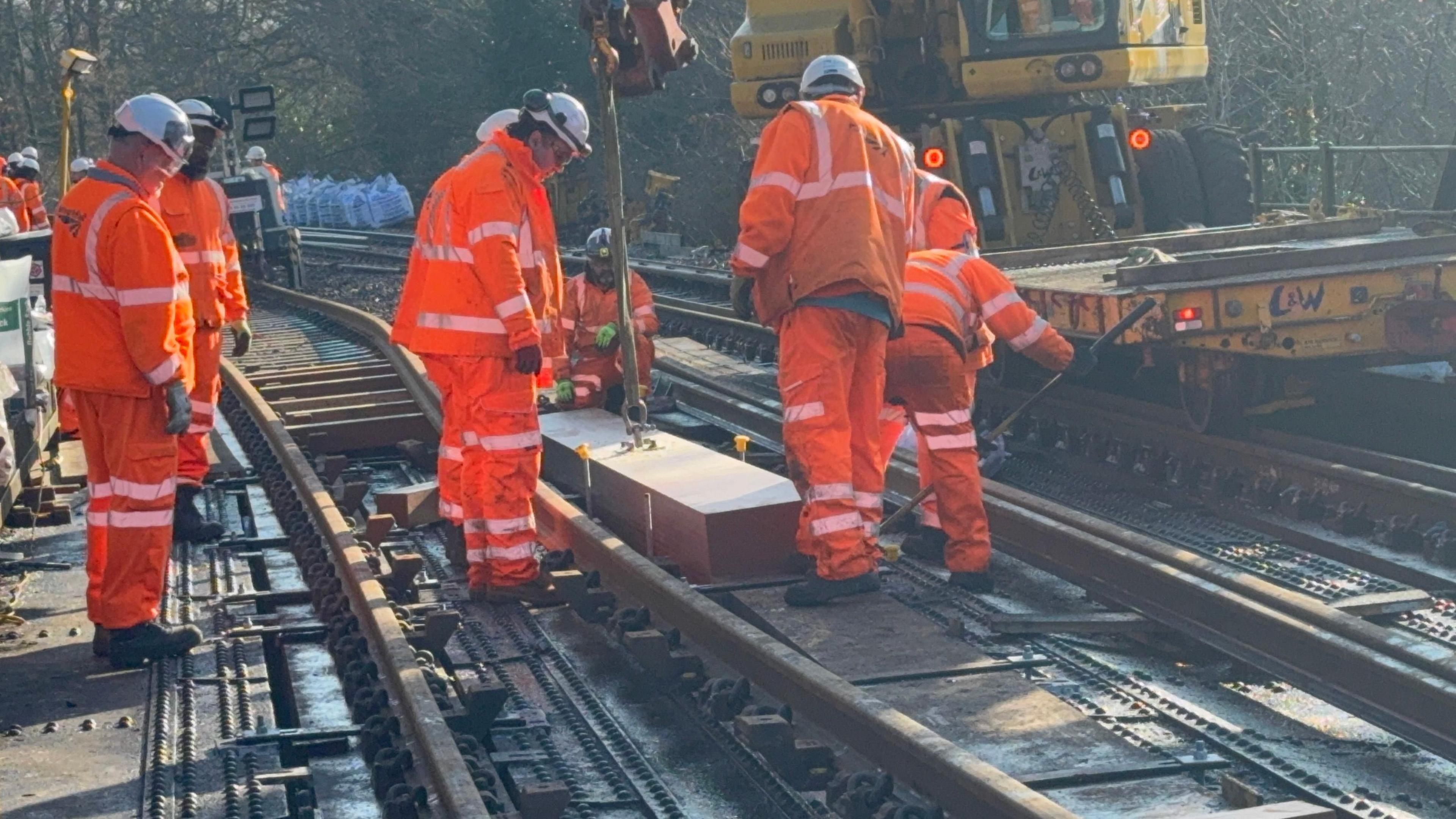 Network Rail workers in orange hi-vis and safety gear stand over a rail, inspecting a piece being added to it.