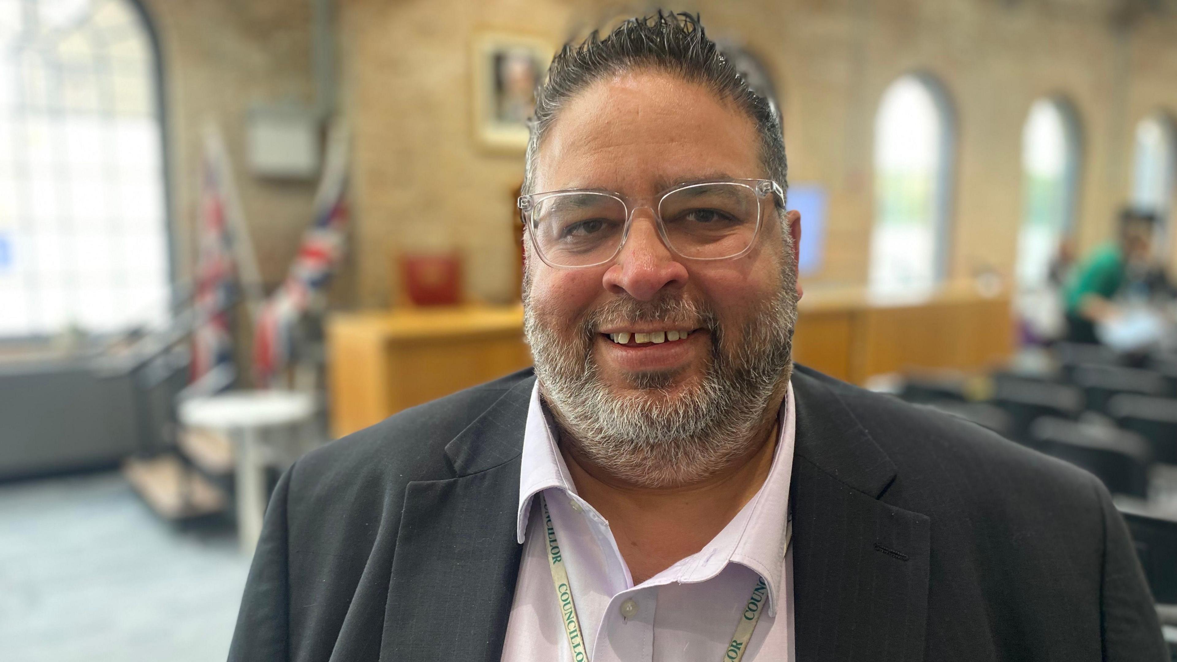 Mohammed Jamil looking into camera. He has short spiky hair, grey beard and is wearing glasses and a dark suit with a light shirt. He is in a council chamber.