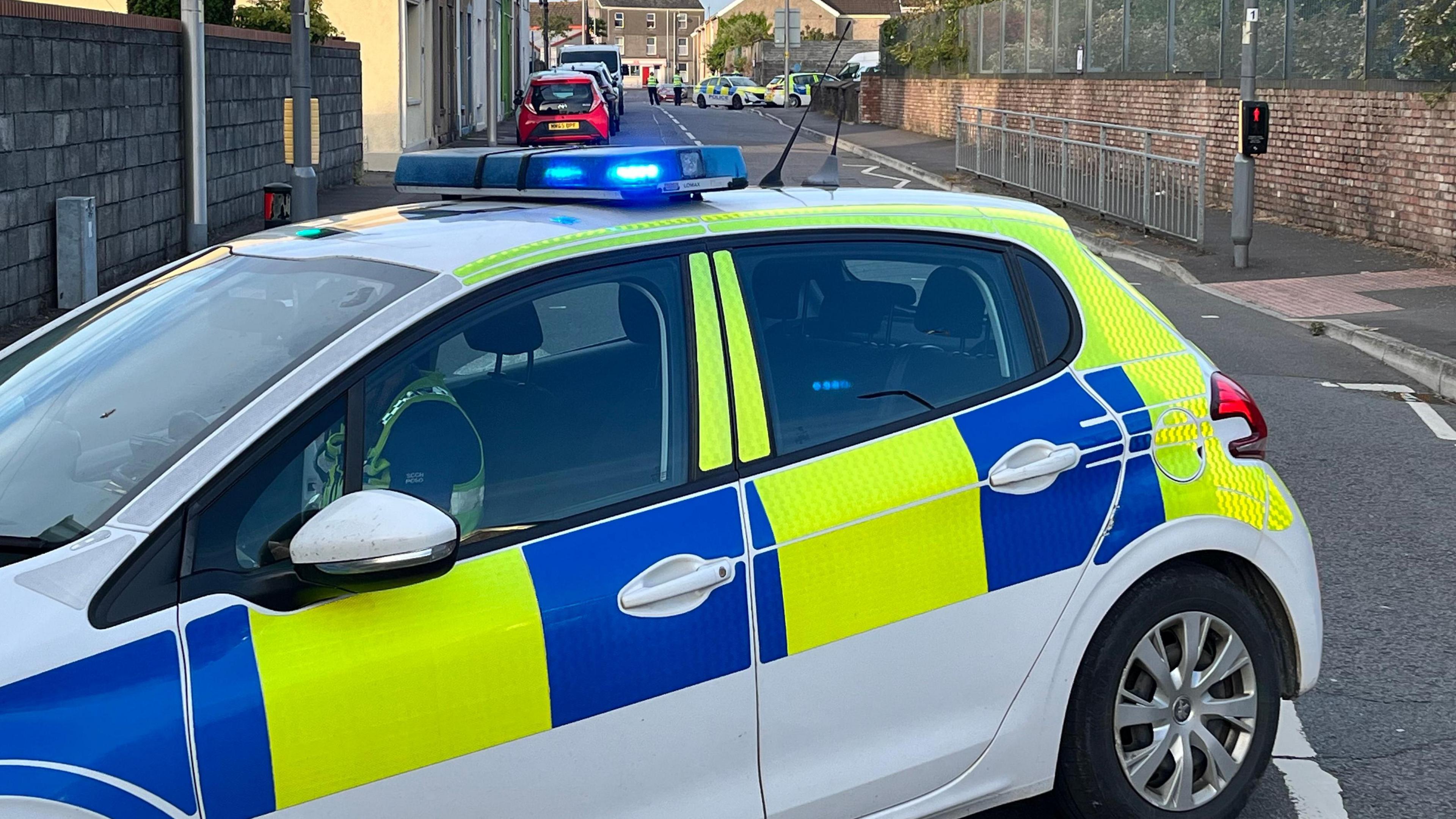 A police car in the forefront, with a road behind and, in the background at the far end of the road, tow more police cars and two uniformed officers.