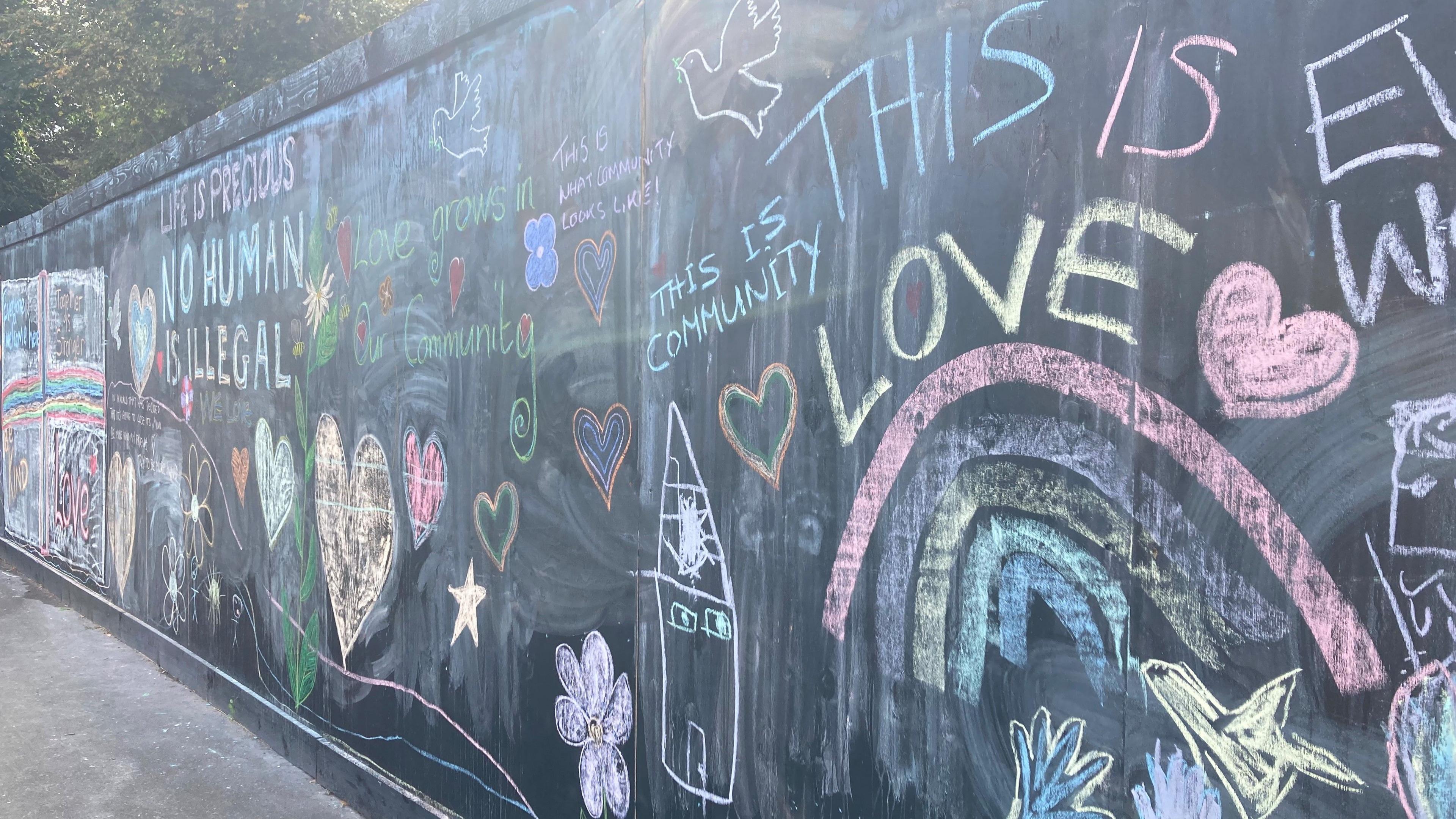 Hoarding on Dereham Road near a construction site. On the hoarding is message of love and support written in chalk. Some of the drawings show love hearts, rainbows and flowers. Messages within the picture includes 'this is community', 'this is love', 'life is previous, no human is illegal'.