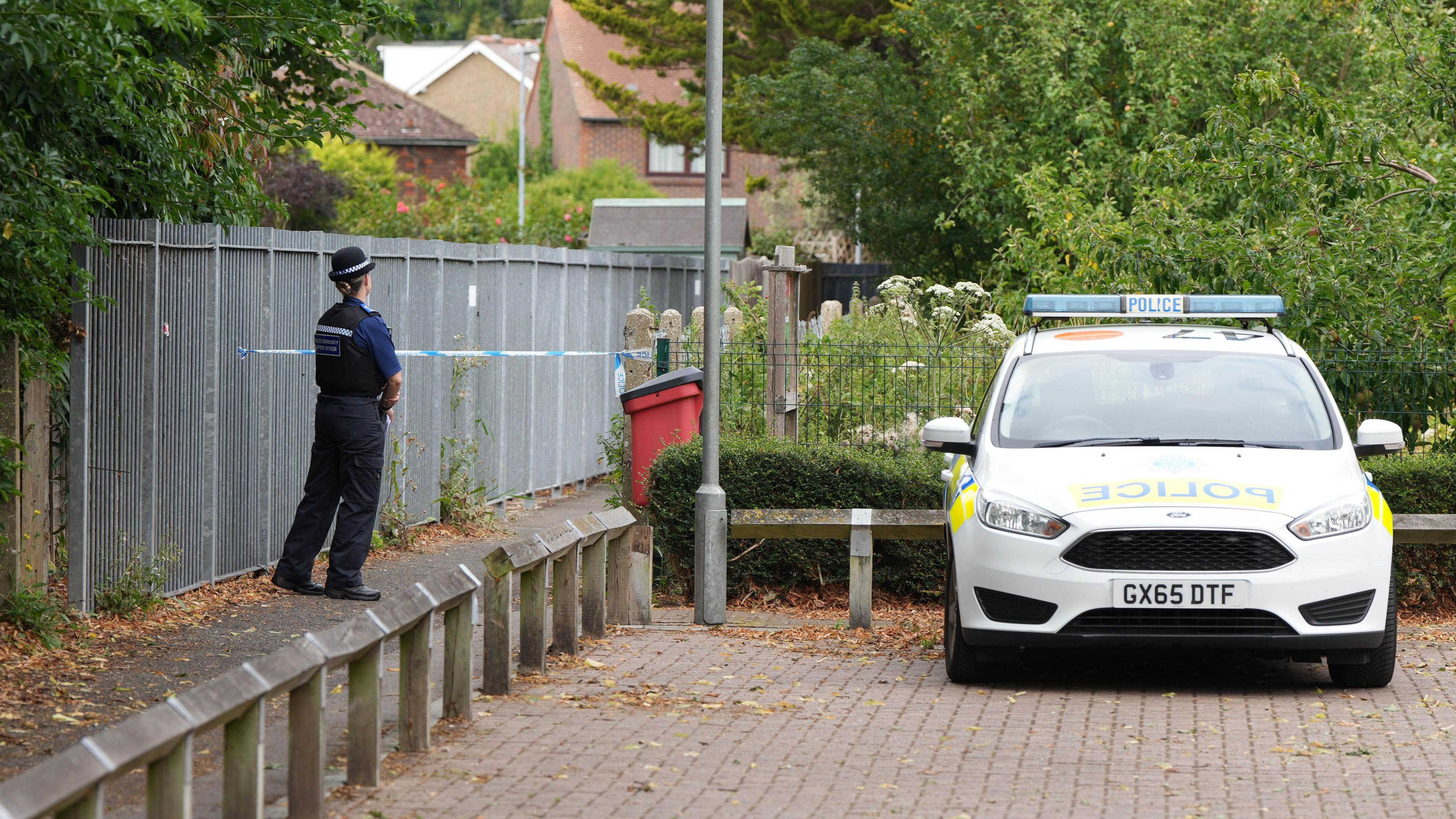 An alleyway taped off with blue and white police tape. A police officer stands at the edge of the tape cordon. A police car is parked next to the cordon.