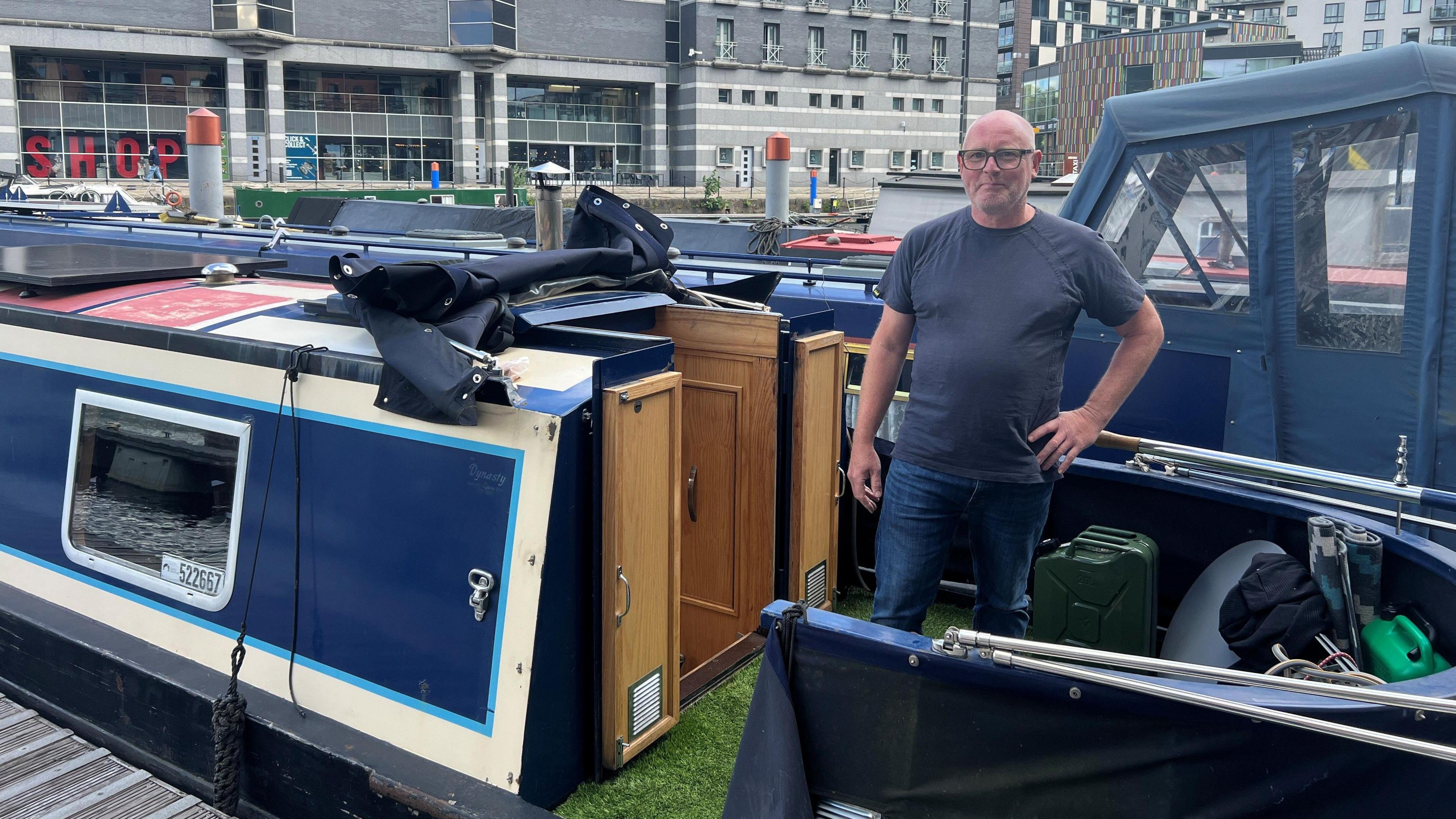 Ken Tracey standing by the doors of his blue and white narrow boat. He is bald, wearing glasses and a blue top and jeans. There are buildings in the background.