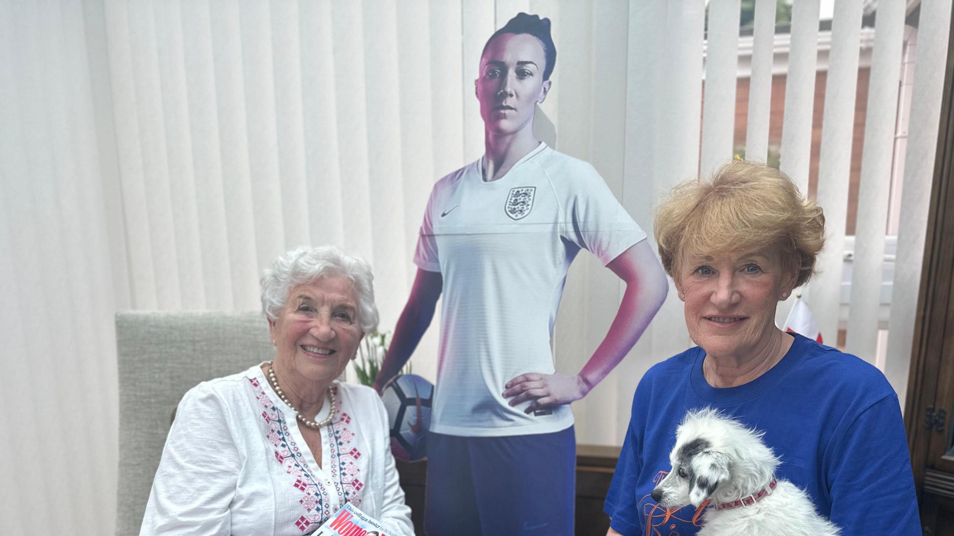 Gloria Stapylton (left) has short white hair and is smiling. She wears a pearl necklace and white cardigan. She is holding a copy of Women's Health magazine, which has a picture of Lucy Bronze on it. Julie Tough (right) has short ginger hair and holds a small white dog and a newspaper. A cardboard cutout of Lucy is behind them.