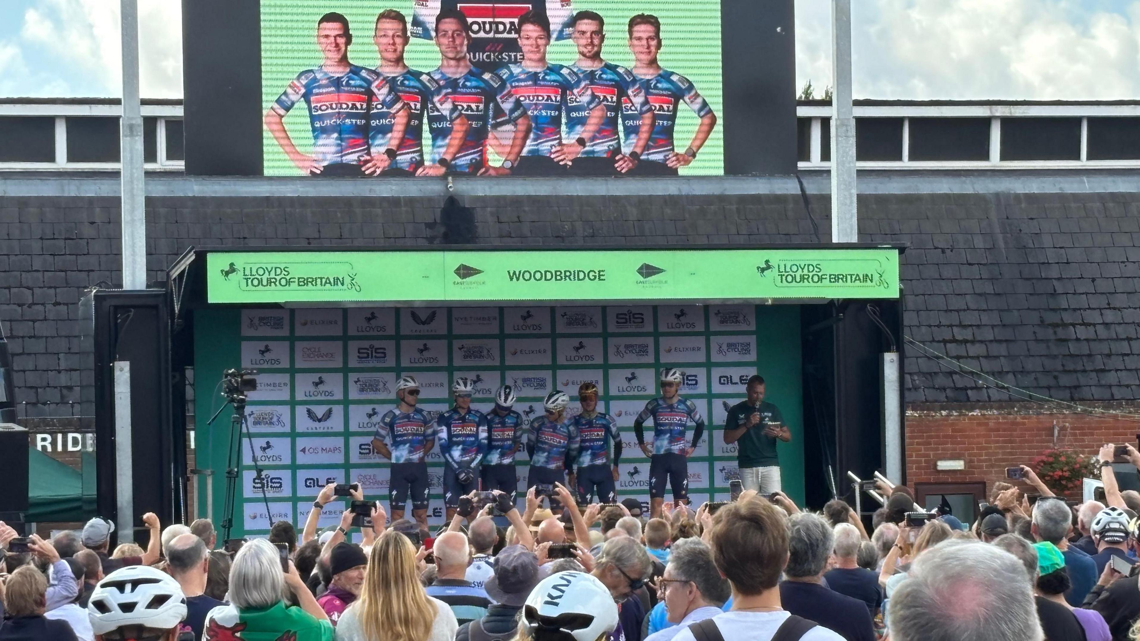 A team of cyclists stand on a stage being presented to a crowd. People watch on with some holding their phones up above the crowd to get a photo.