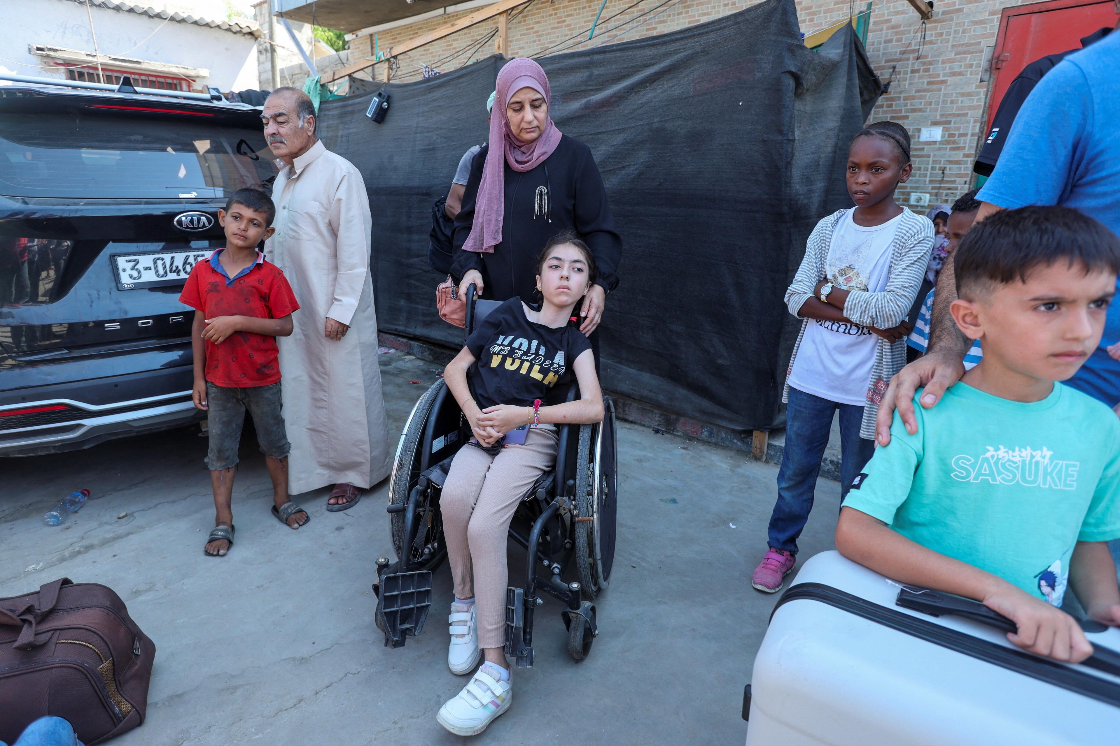 Shatha Abusalim, wearing a dress and headscarf, stands behind her daughter, Lamis, wearing a t-shirt and trousers, as the girl sits in a wheelchair waiting on the street to be evacuated. Other children and adults stand near them, one with a suitcase