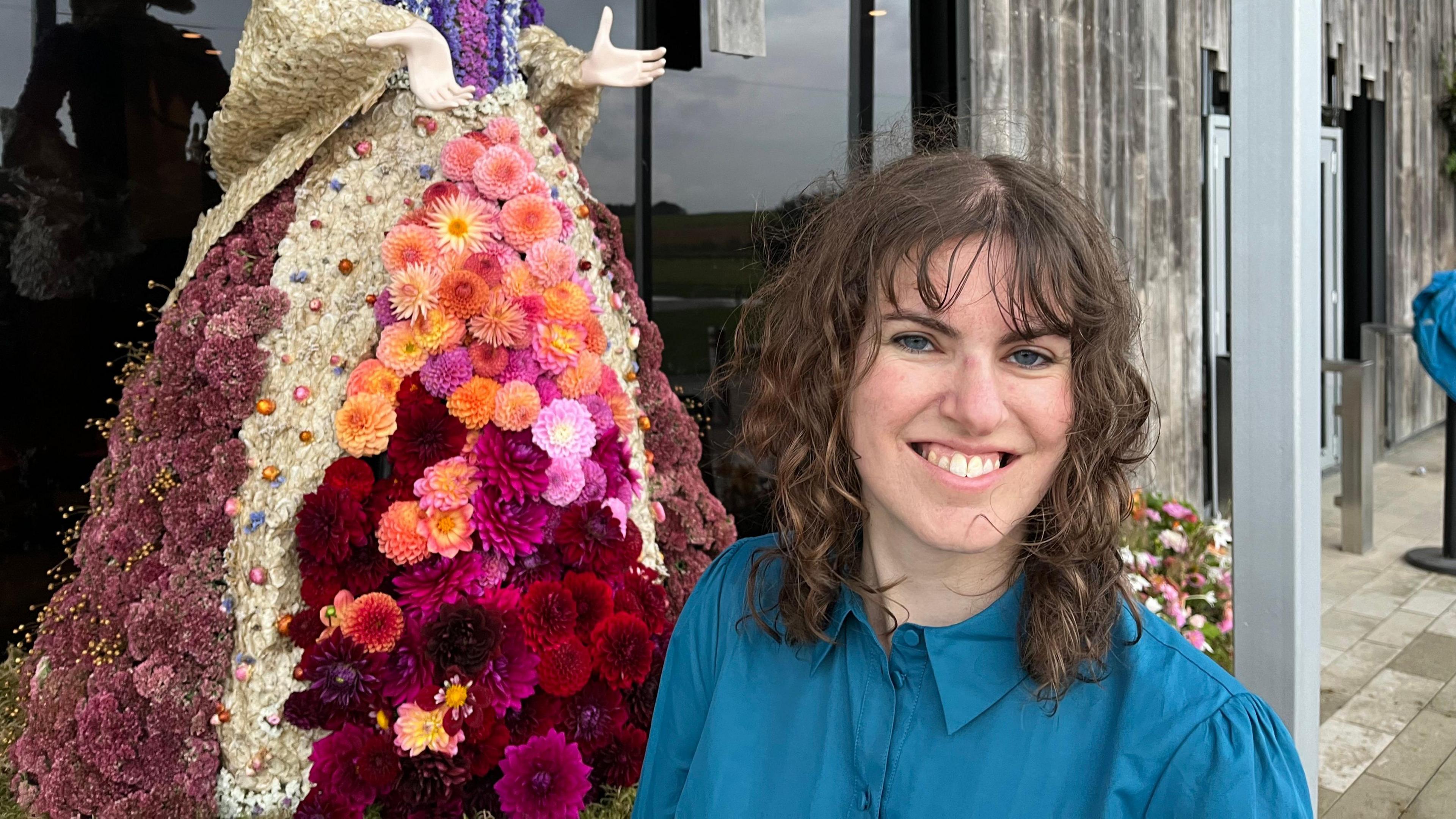 Emily Parker in a blue blouse with curly shoulder-length brown hair and a fringe. She is standing beside the skirt section of a dahlia sculpture. There are pleated layers of different types and colours of dahlias. 