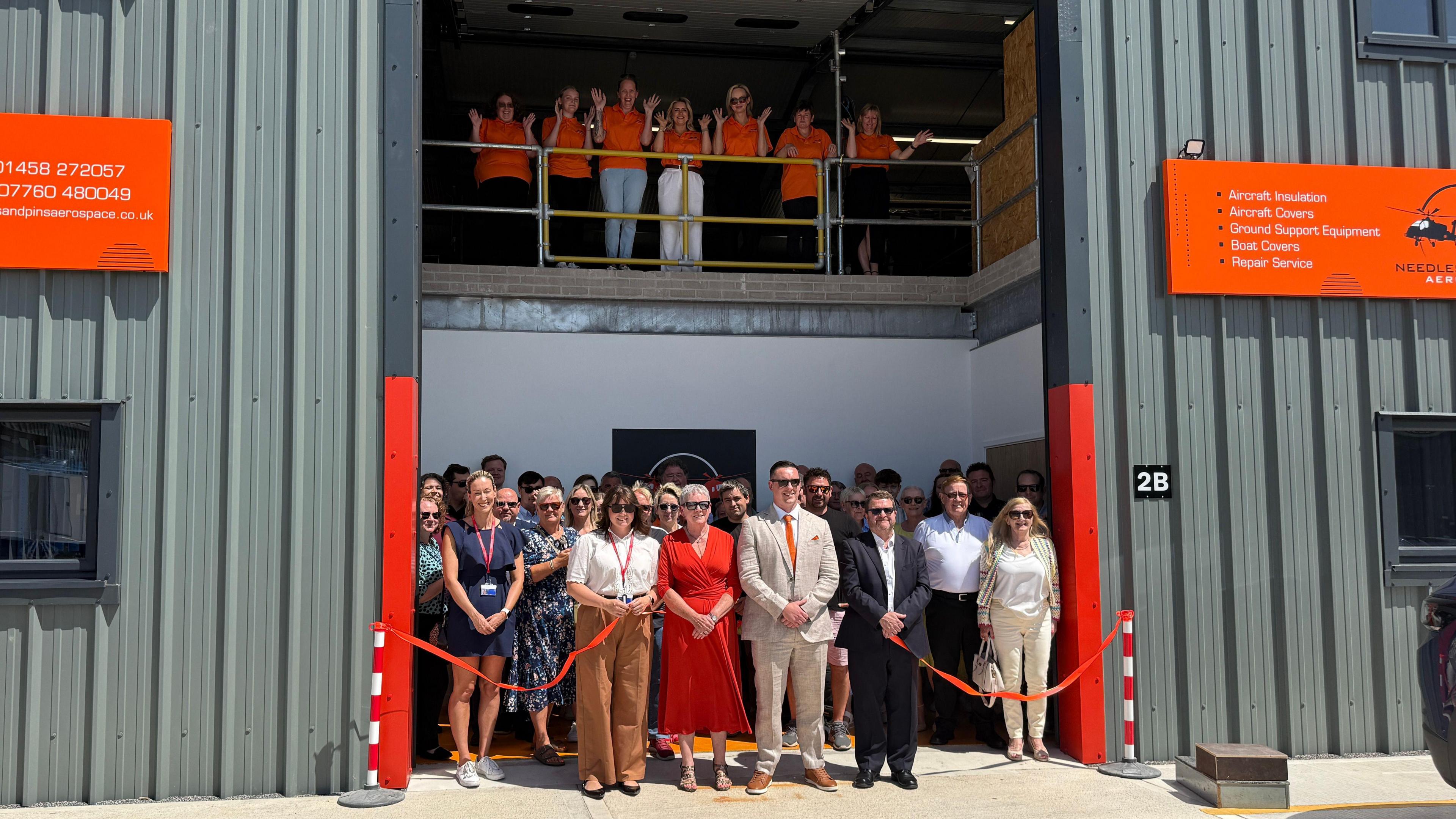 A group of people from the Needles and Pins team standing behind red ribbon at the opening of an industrial steel building.