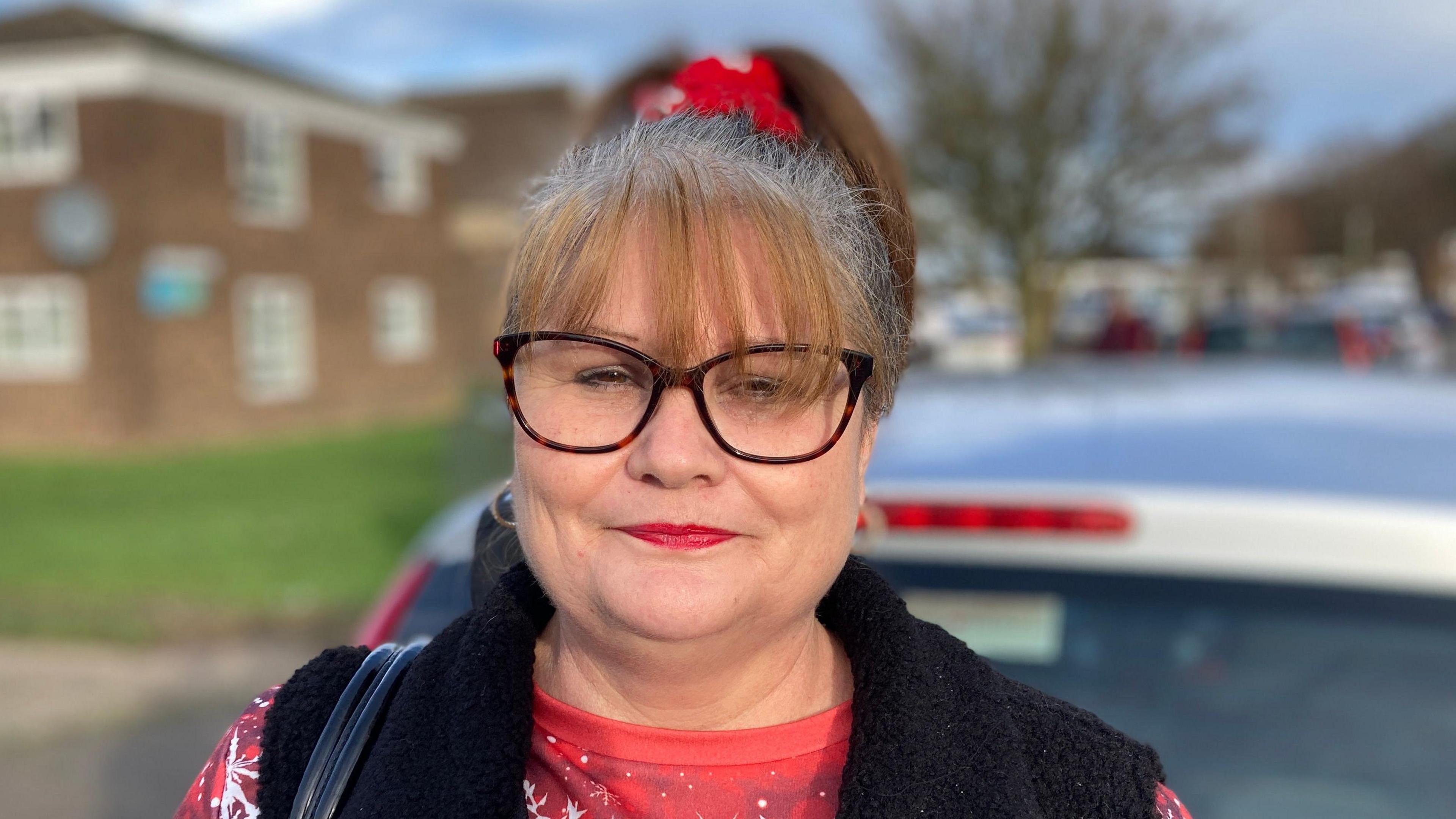 Melanie Hullott. She is wearing a red Christmas jumper and a black gilet. She has glasses. She is standing outside.