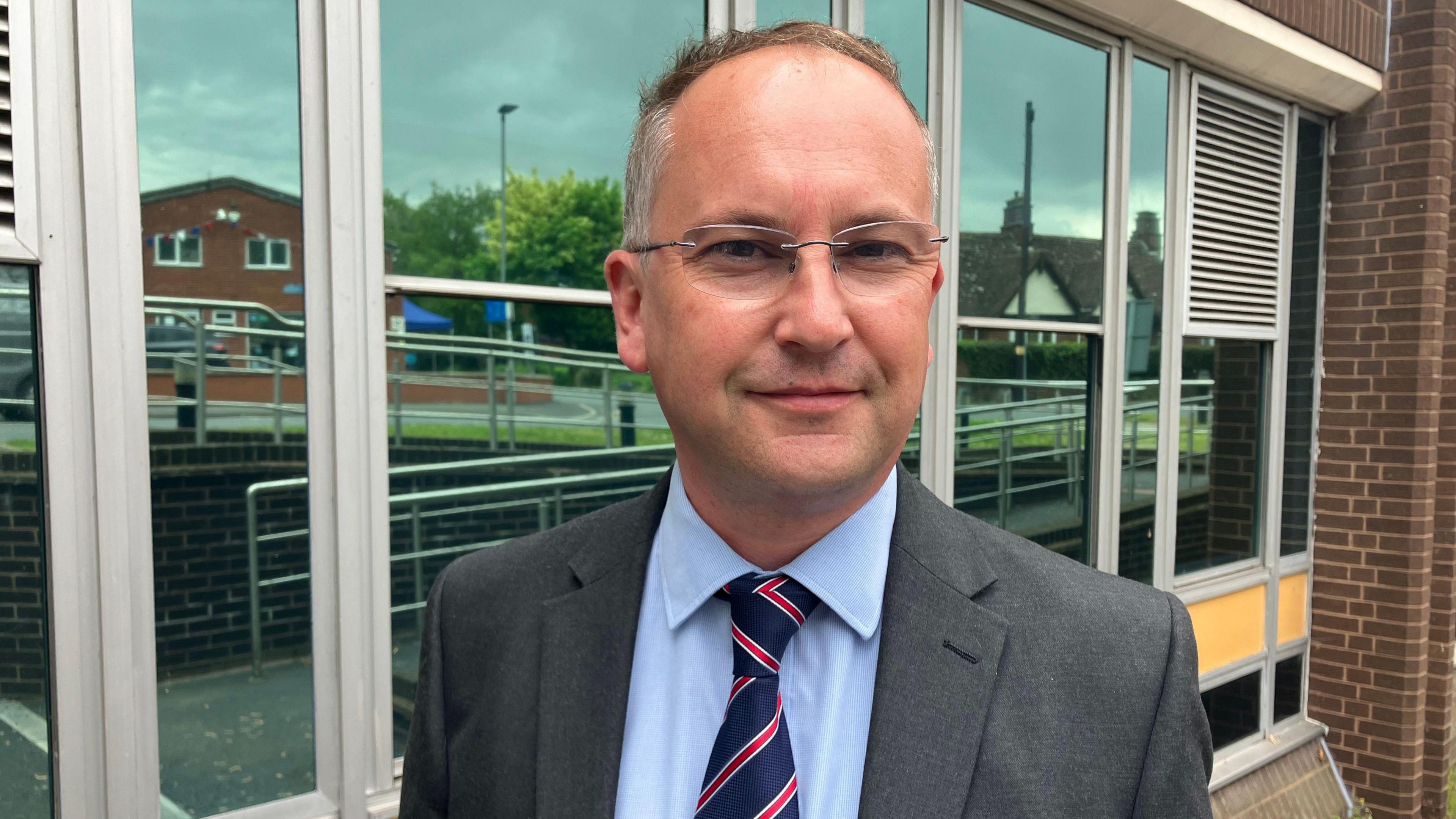A man wearing a grey suit with a blue shirt and a red and blue tie, stood behind a modern glass-fronted building.
