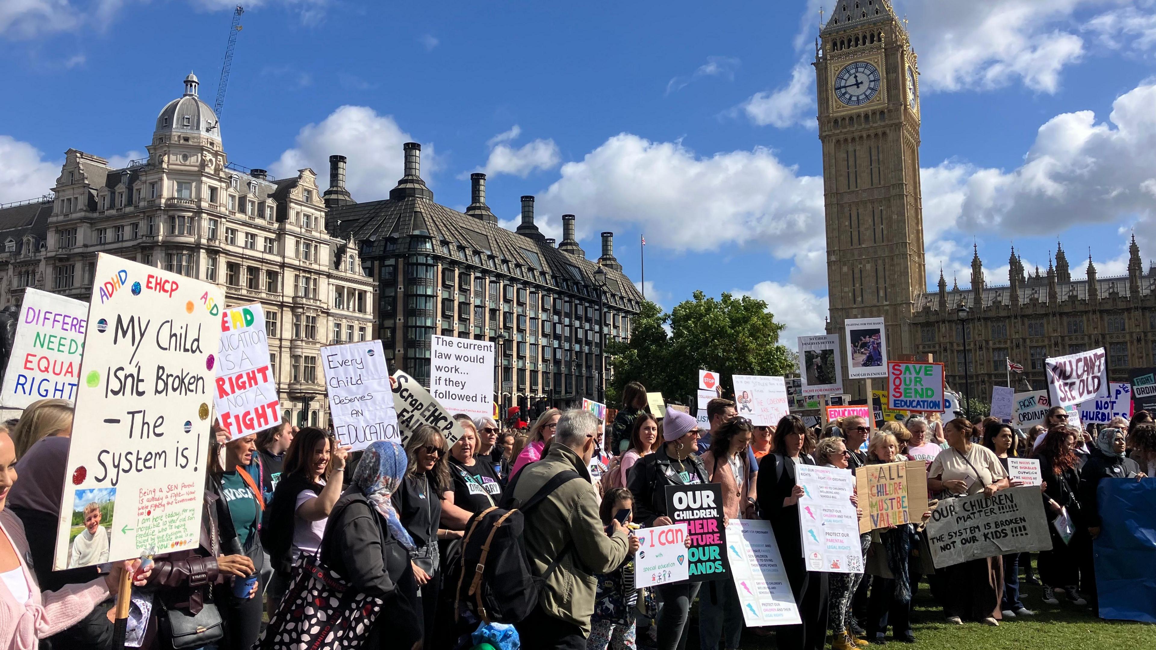 Dozens of parents stand on a sunny Parliament Square with Big Ben in the background holding signs calling for the rights of their children with special educational needs to be protected. The most prominent sign says 'My child isn't broken -the system is."