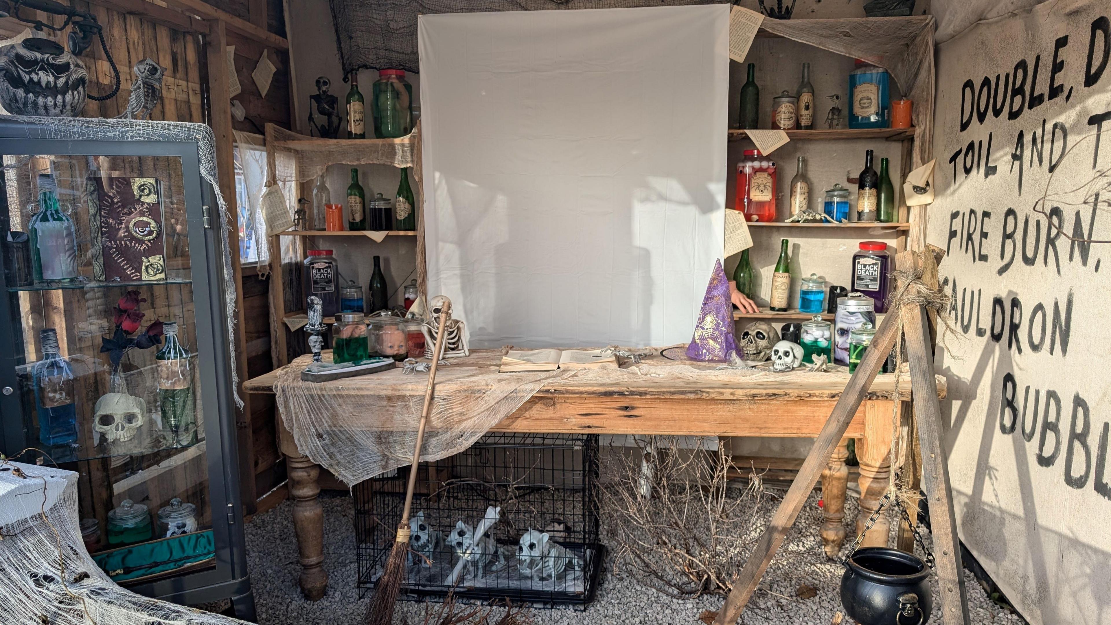 A spooky workshop with a class cabinet on the left with skills and potion bottles. Shelves at the back also have potion bottles, with a wooden table in front.