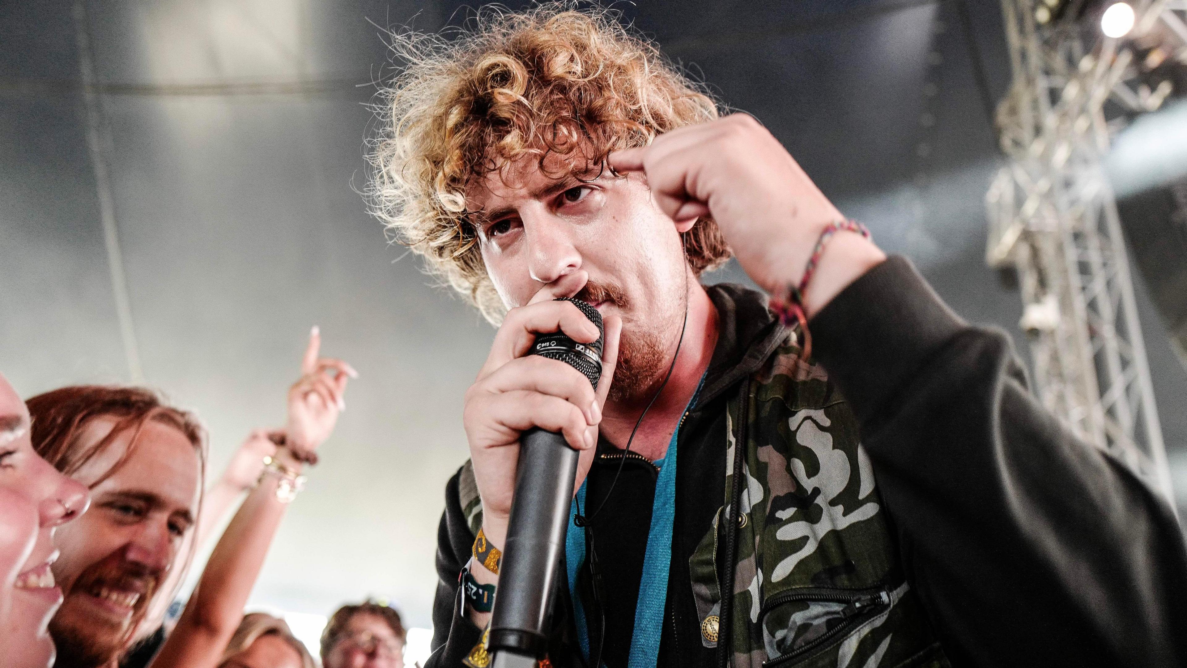 Joe Love, a man with curly blond hair, holding a microphone with one hand and gesturing with another. He is standing very close to the front row of a crowd watching him. The background includes stage equipment including a silver metal structure.