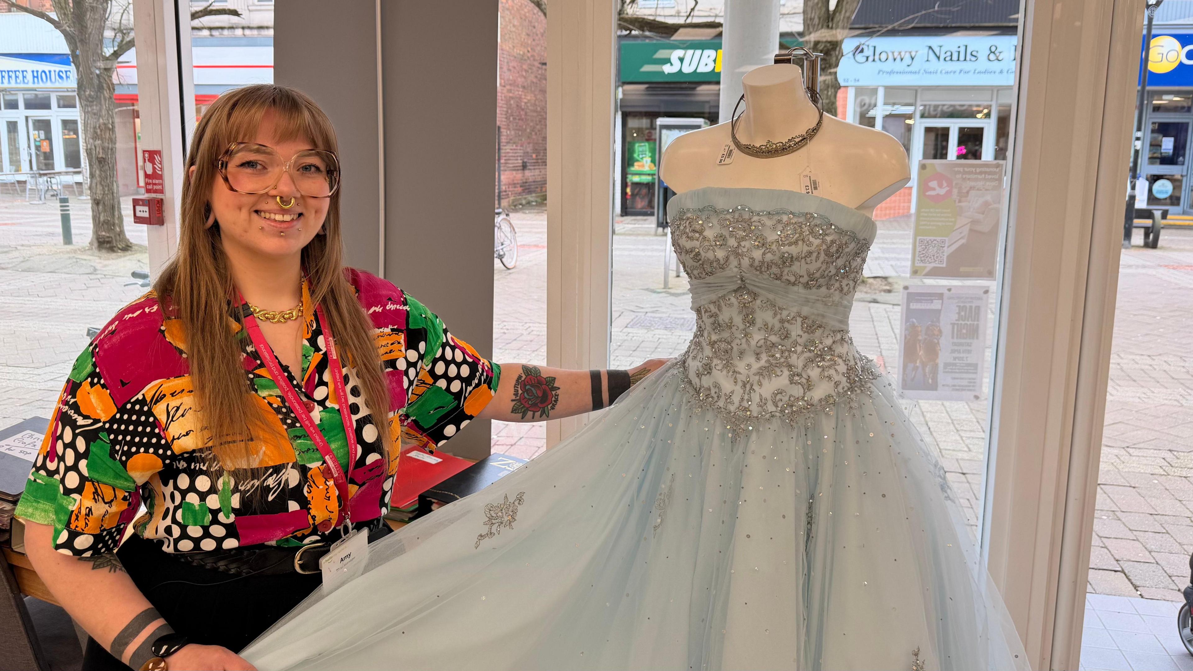 A woman smiles as she holds a princess dress, which is baby blue with glittery embellishments, on a mannequin in a shop window, with a high street outside. The woman wears large glasses and a bold-patterned multi-coloured shirt. She  has prominent facial piercings. 