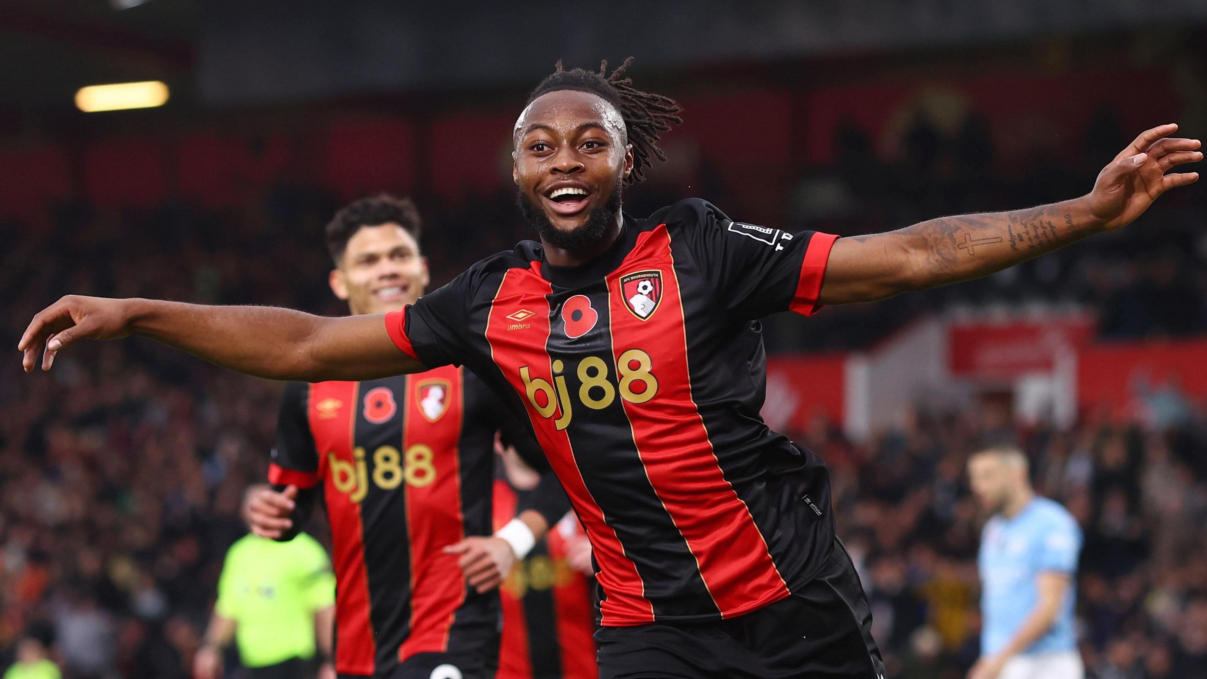 Antoine Semenyo, wearing a red and black striped Bournemouth strip, stretches his arms out wide and smiles while celebrating a goal against Manchester City