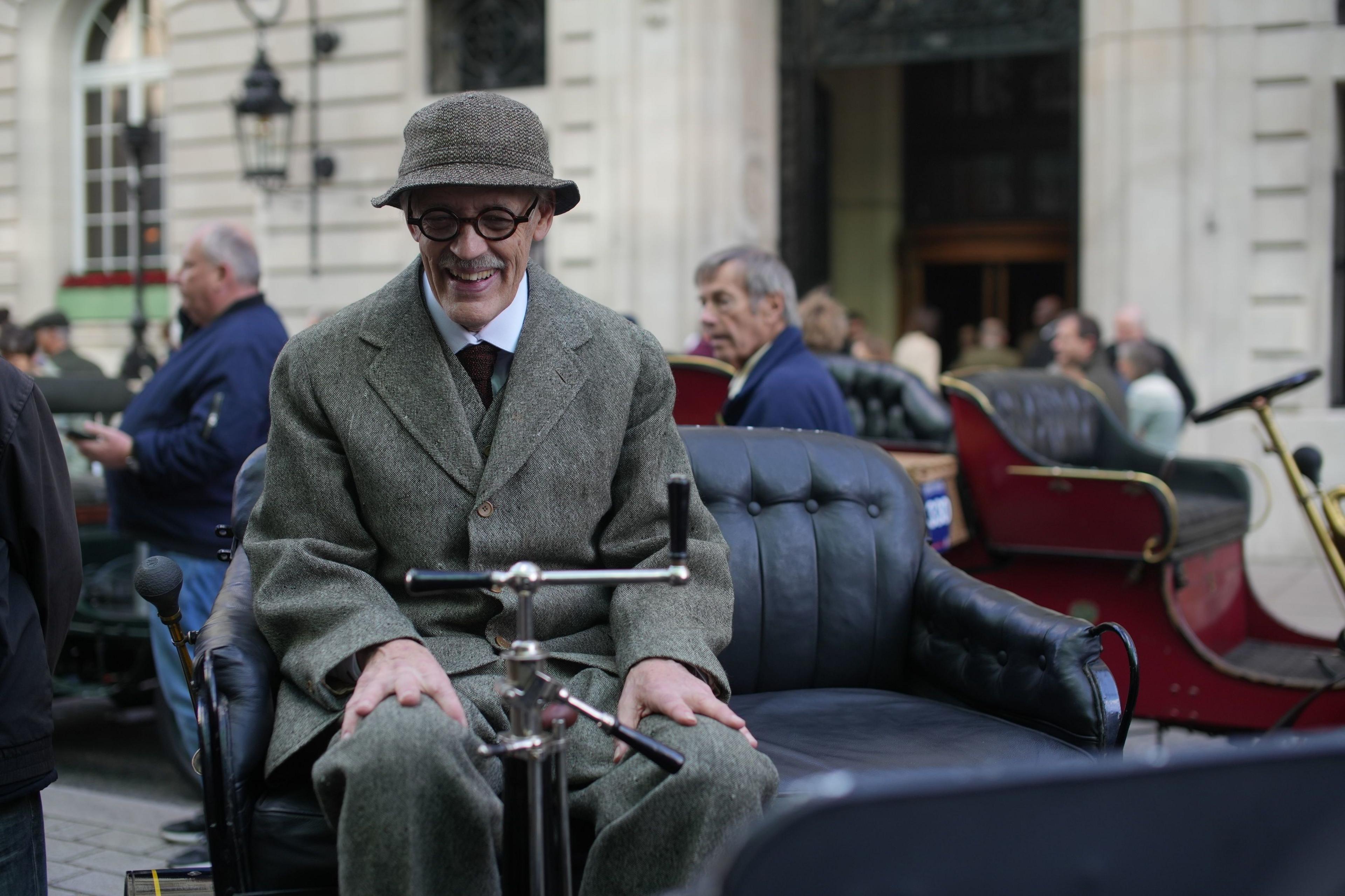 A man in an old-fashioned suit, with a hat and glasses, sits on an old car