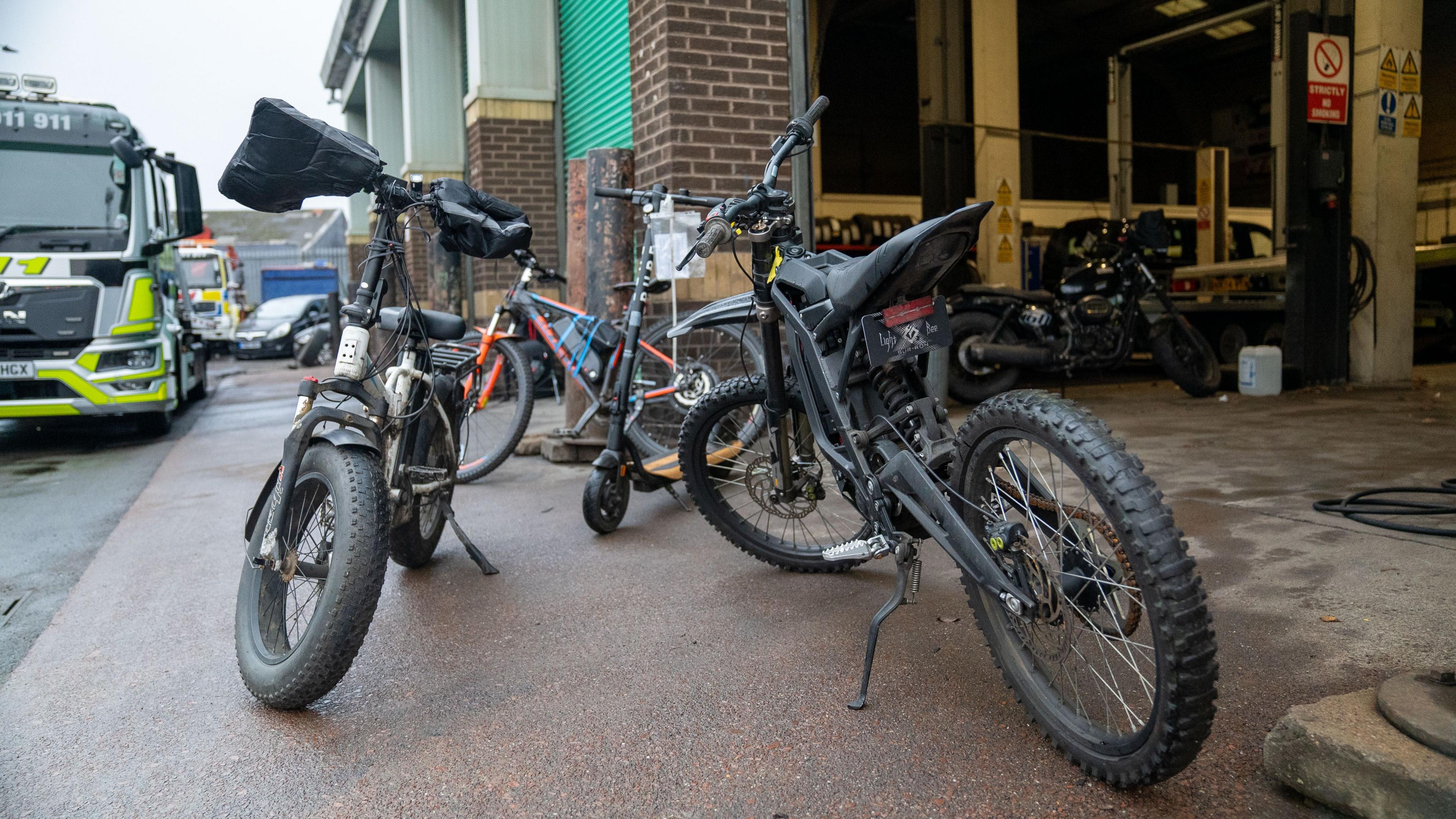 An black e-scooter, an electric motorbike and a white pedal bike with thick wheels and a white body pictured in a scrap yard.