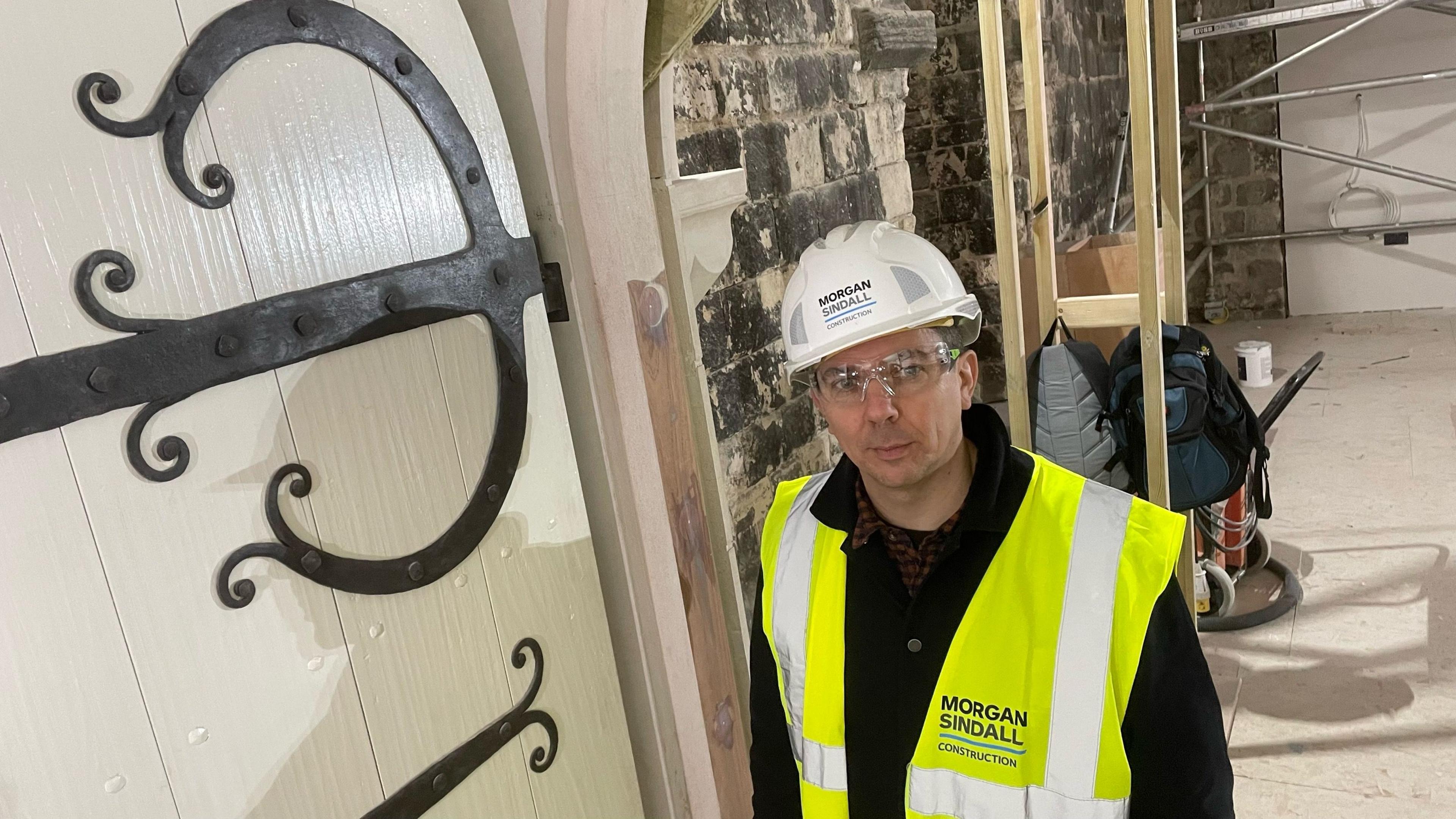 James Spedding is wearing a high-vis jacket and a white helmet stood next to a large wooden door with steel braces and decorations