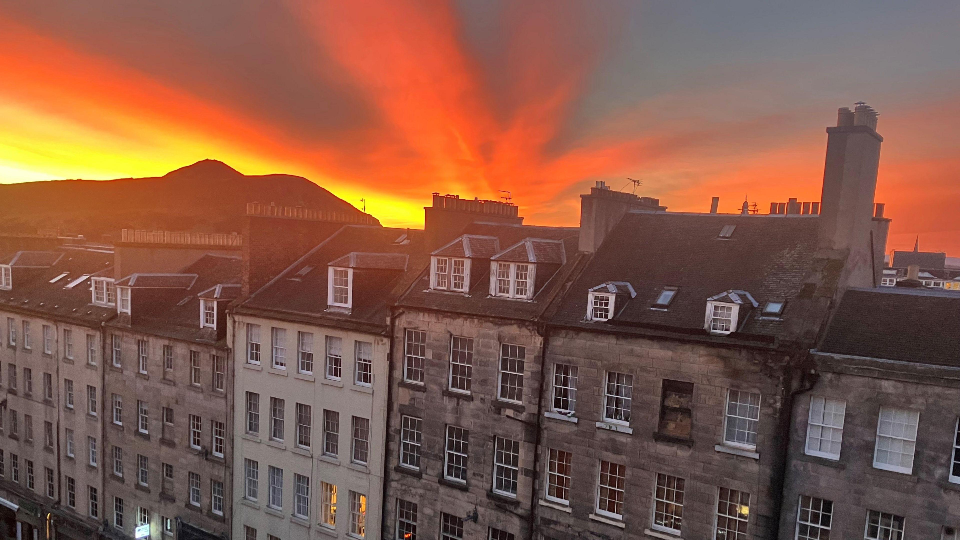 There are tenement buildings in the foreground. In the background is the silhouette of Arthur' Seat. The sky is orange, red and yellow.
