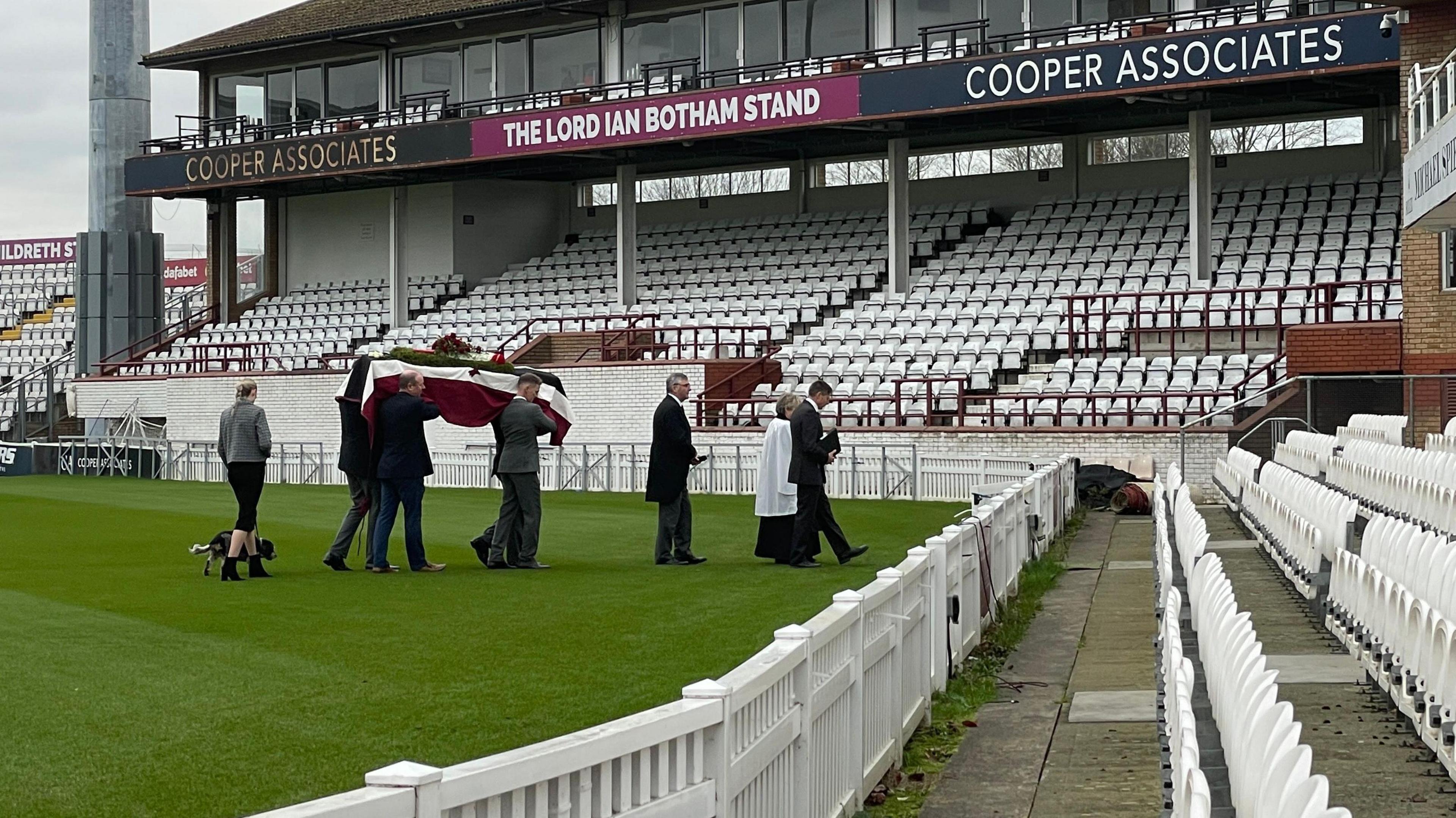 Mary Elworthy-Coggan's coffin is being carried across a cricket pitch towards the stands.