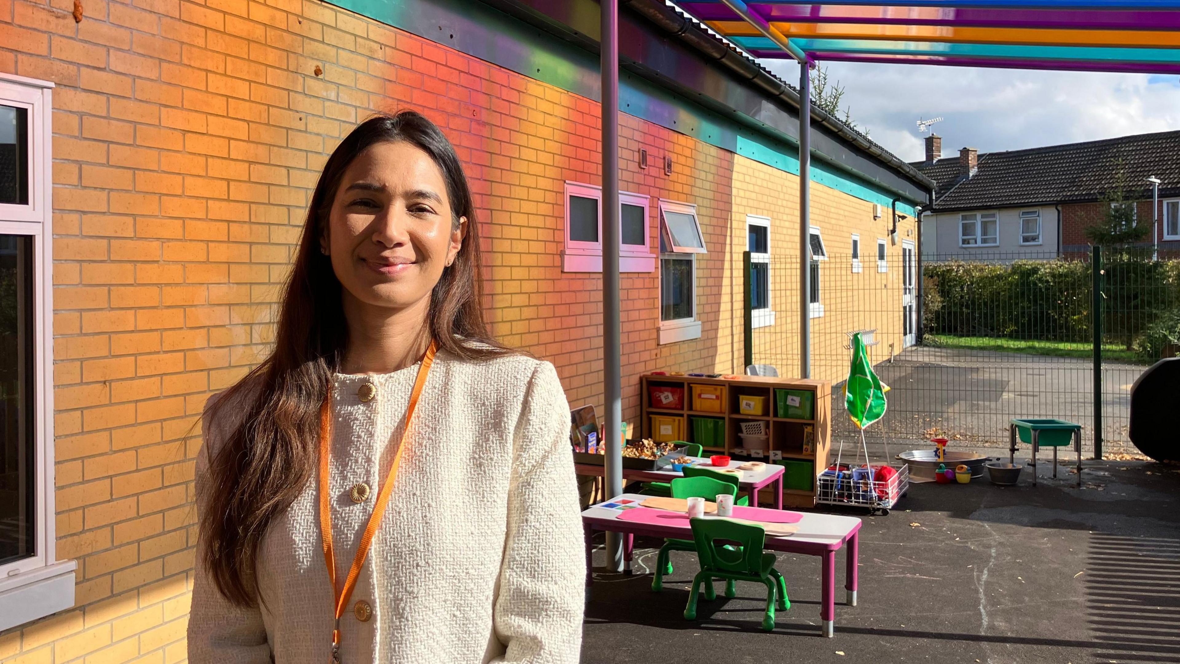 Adorabelle Amaral-Shaikh wearing a white jumper and orange lanyard with a playground setting behind her with chairs, tables and drawers with a rainbow outdoor roof above her.