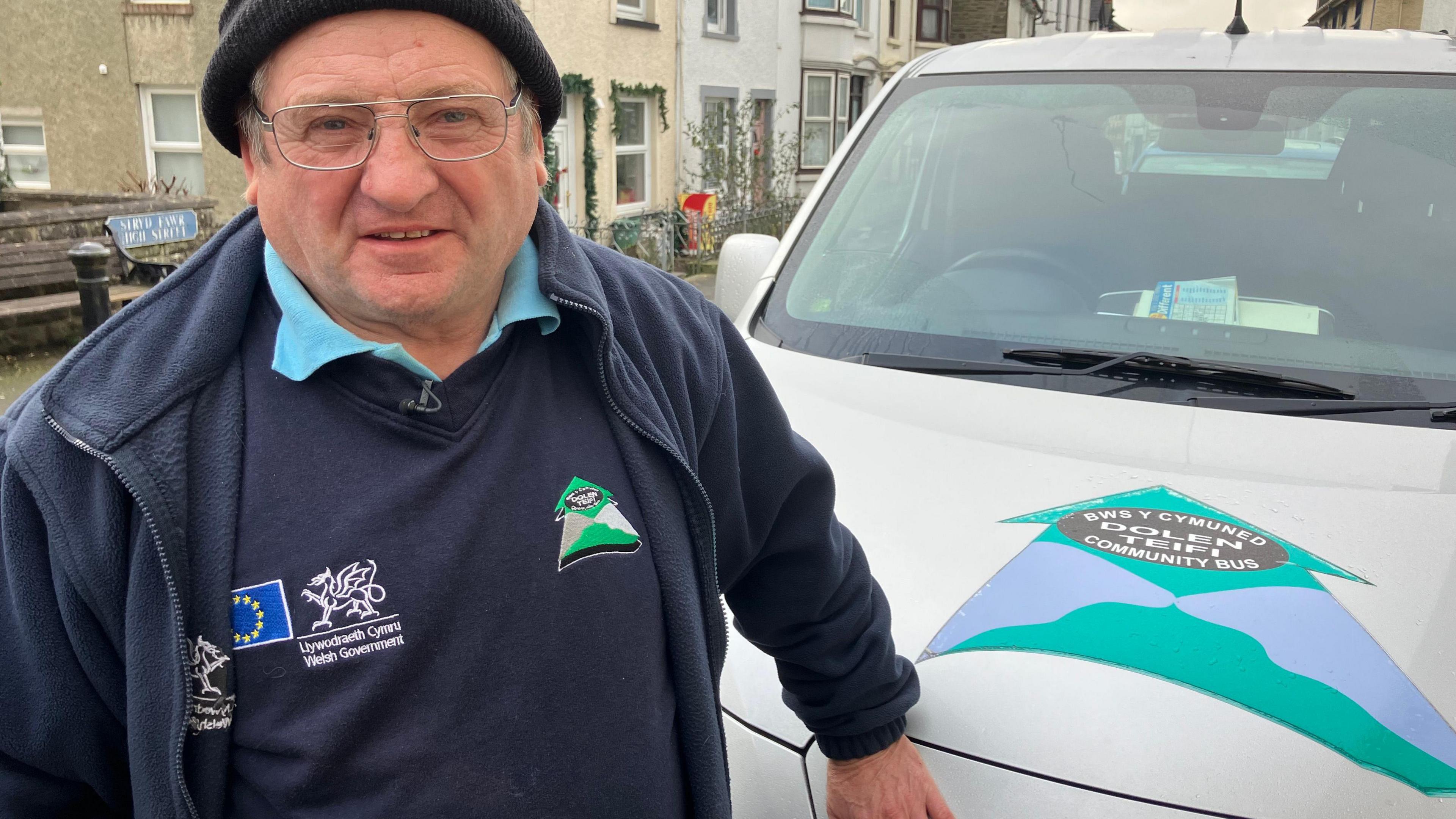 John Hands, a volunteer driver for Dolen Teifi, wearing a beanie and a blue fleece over a blue shirt and jumper, smiles at the camera in front of the community transport vehicle he uses to ferry people around. The vehicle carries the Dolen Teifi logo, along with Community Bus, and Bws y Cymuned