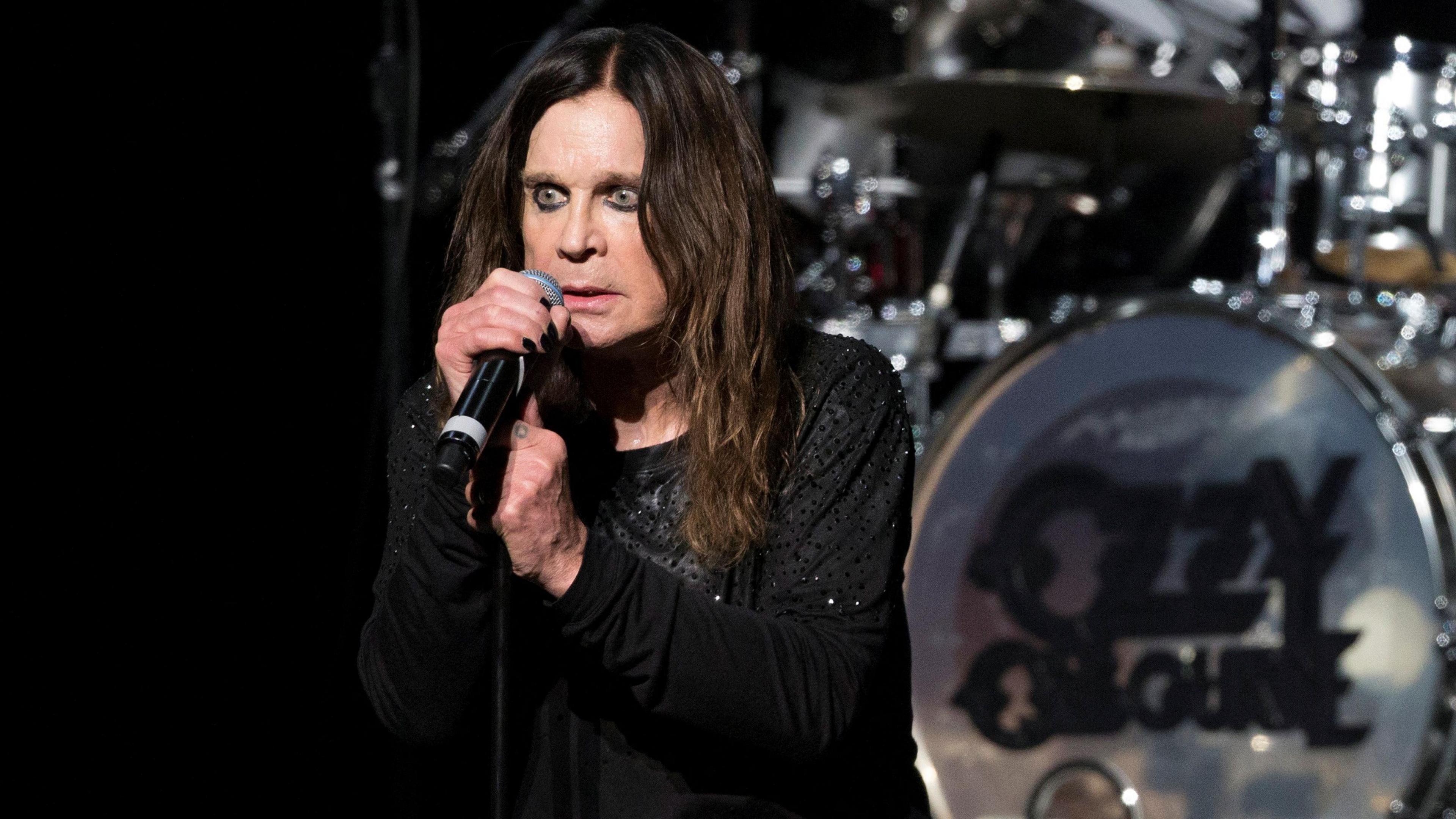 Ozzy Osbourne - a man with long dark hair and wearing all black - stands with his hands on a microphone and stand with a wide-eyed expression on his face. Behind him is a drumkit that says "Ozzy Osbourne".