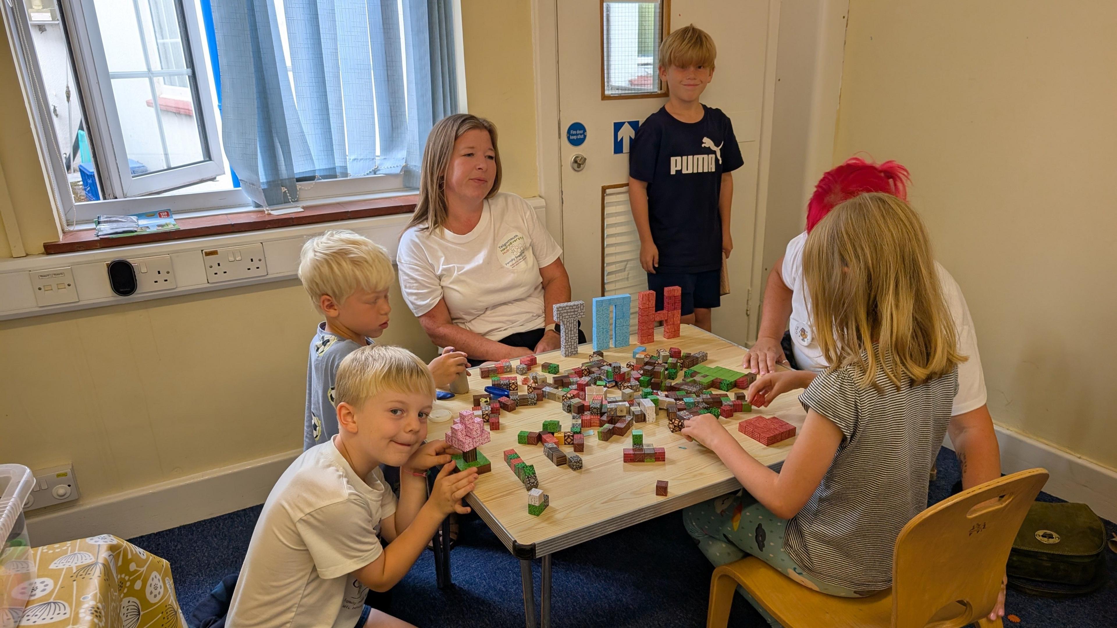 Four children and two adults are sitting around a table playing with building blocks. Two of them are cheekily looking at the camera. 