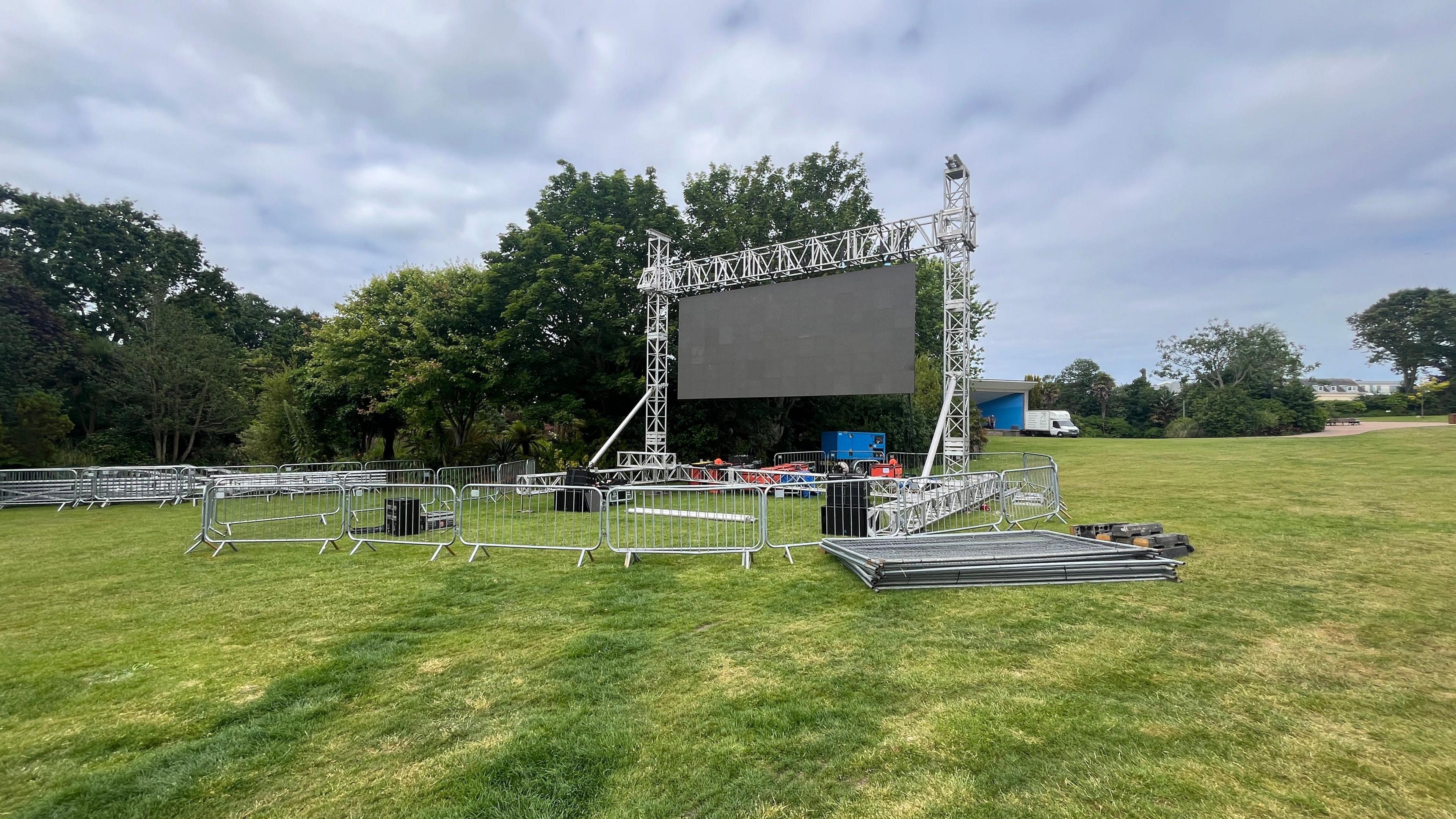 A large LED screen in a park. There is fencing around the screen and trees around the park. 