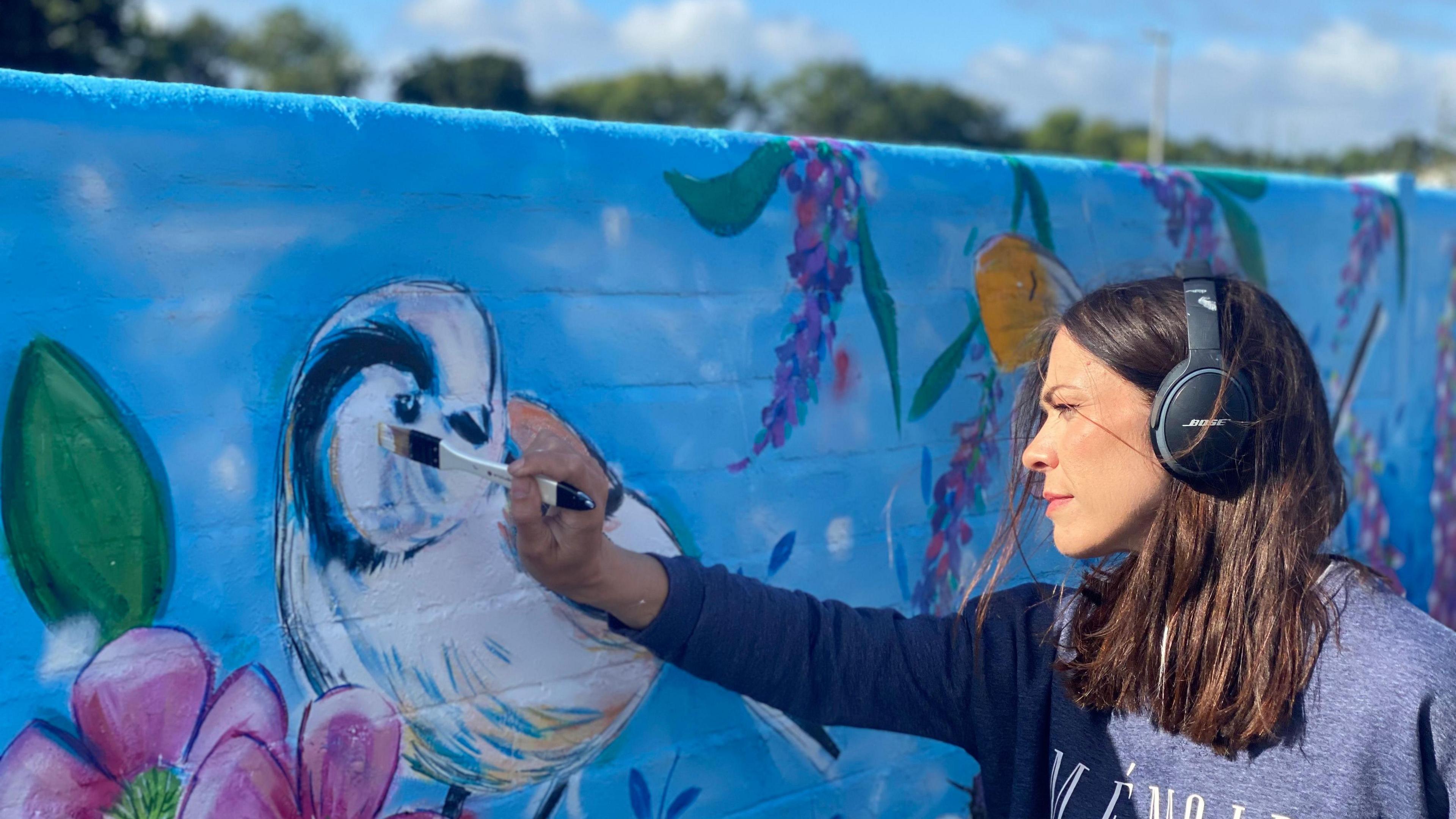 Side view of Chloe Farrant painting the wall. She is wearing the same clothes and is painting a bird with white, yellow and blue plumage.

