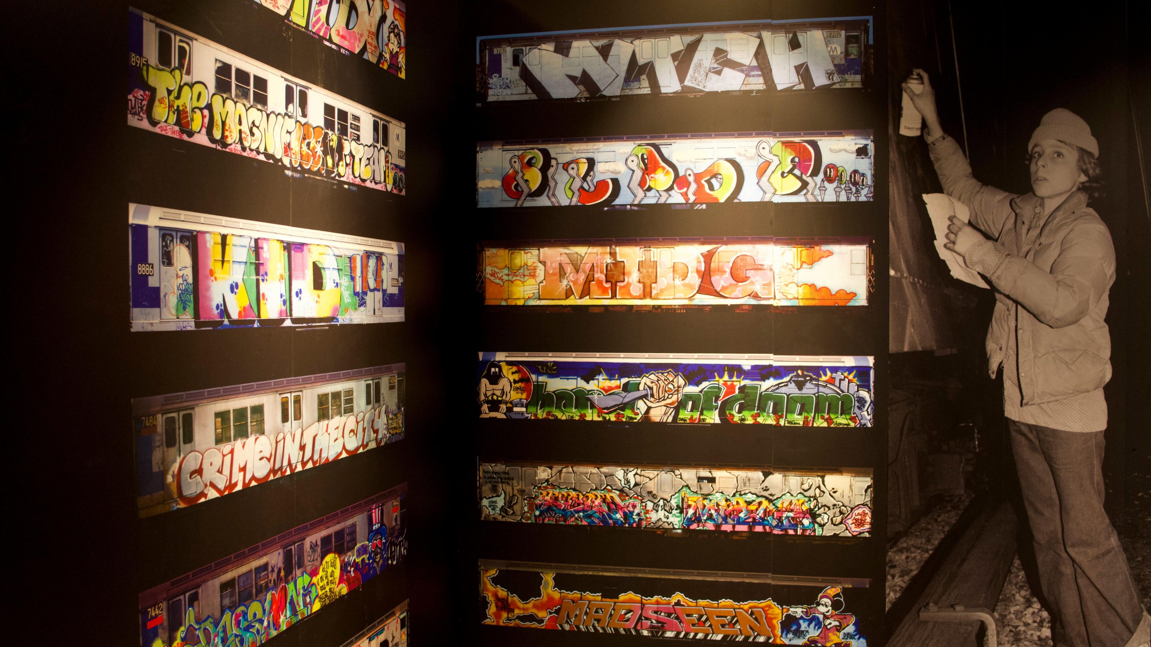 A dozen images of subway trains line a wall at the exhibition, and are next to an image of a teenager in a woolly hat spray-painting a train.