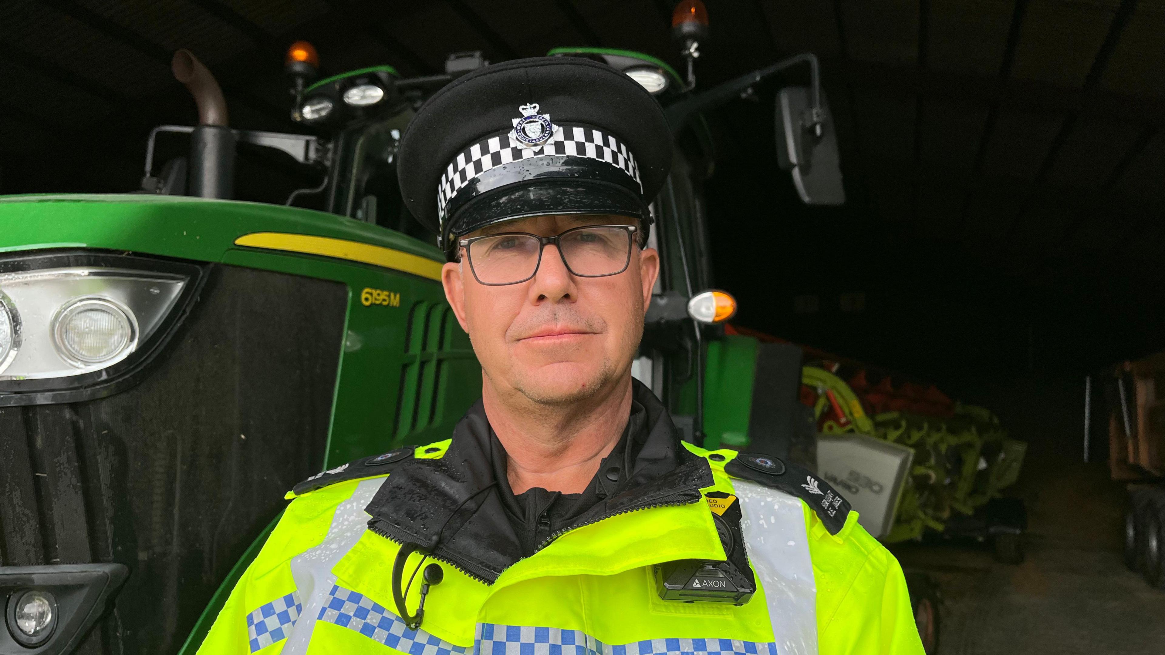 Sgt Jones looking into the camera with a serious expression. He's wearing a fluorescent yellow jacket with blue and white police checks in a stripe across the chest. He's also wearing a black peaked police officer's hat. He's stood in front of a large green John Deere tractor in a farmyard barn. 