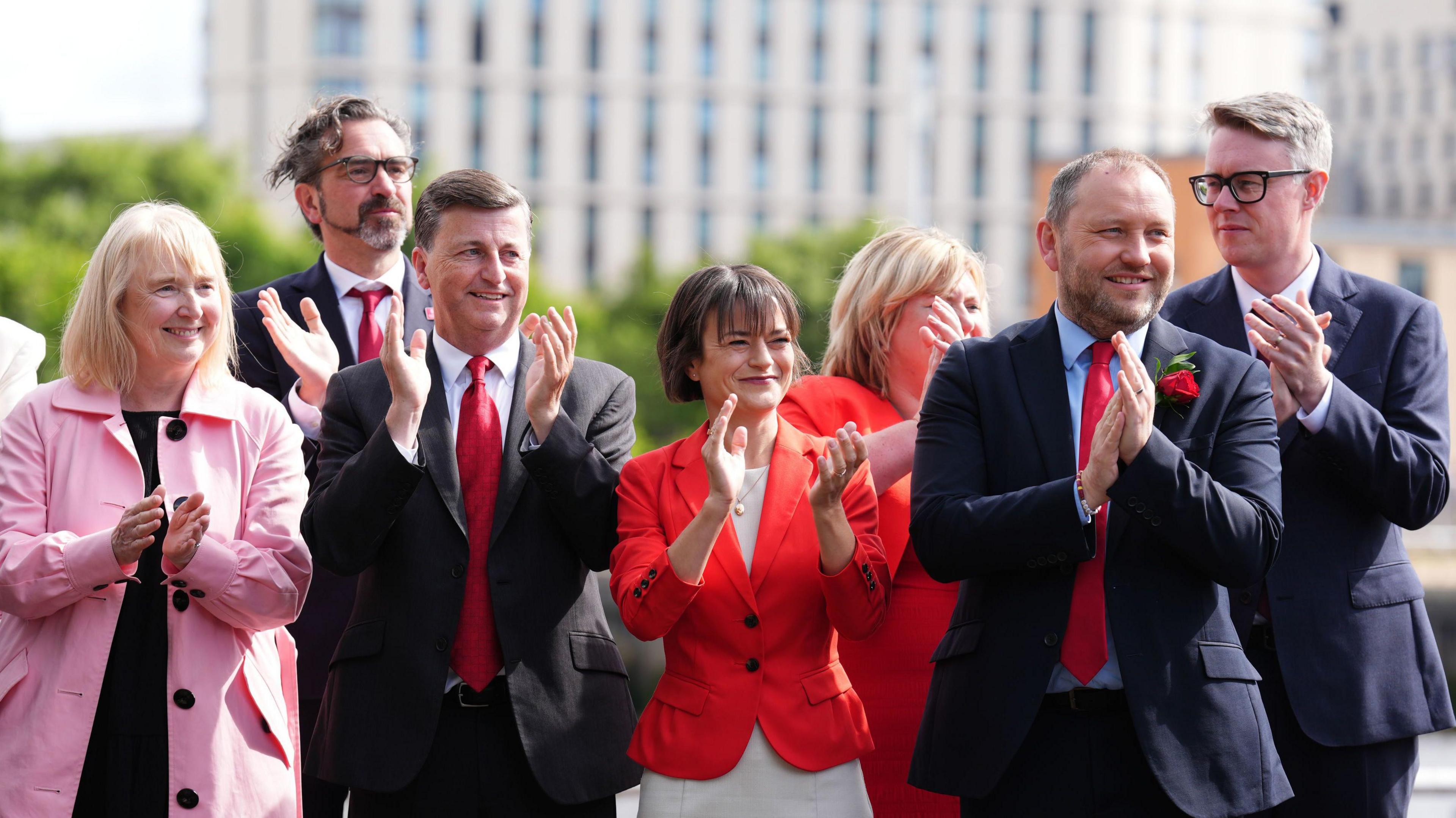 A picture from 2024 of new Labour MPs arriving at Westminster - they include Ian Murray (a man with light hair and a beard) and Douglas Alexander (a man with dark hair - both are wearing bright red ties) - along with several others, all clapping and smiling