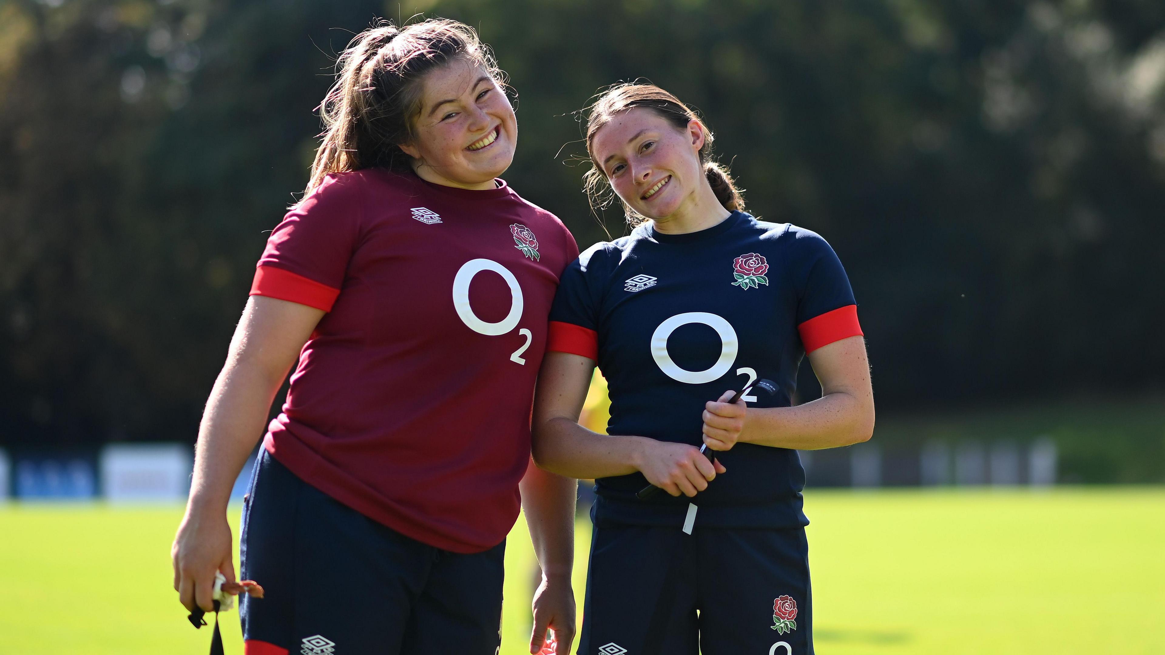 Maud Muir smiling for the camera with another woman player. They are both wearing the Red Roses kits. They are outside on a playing field and it is a sunny day.