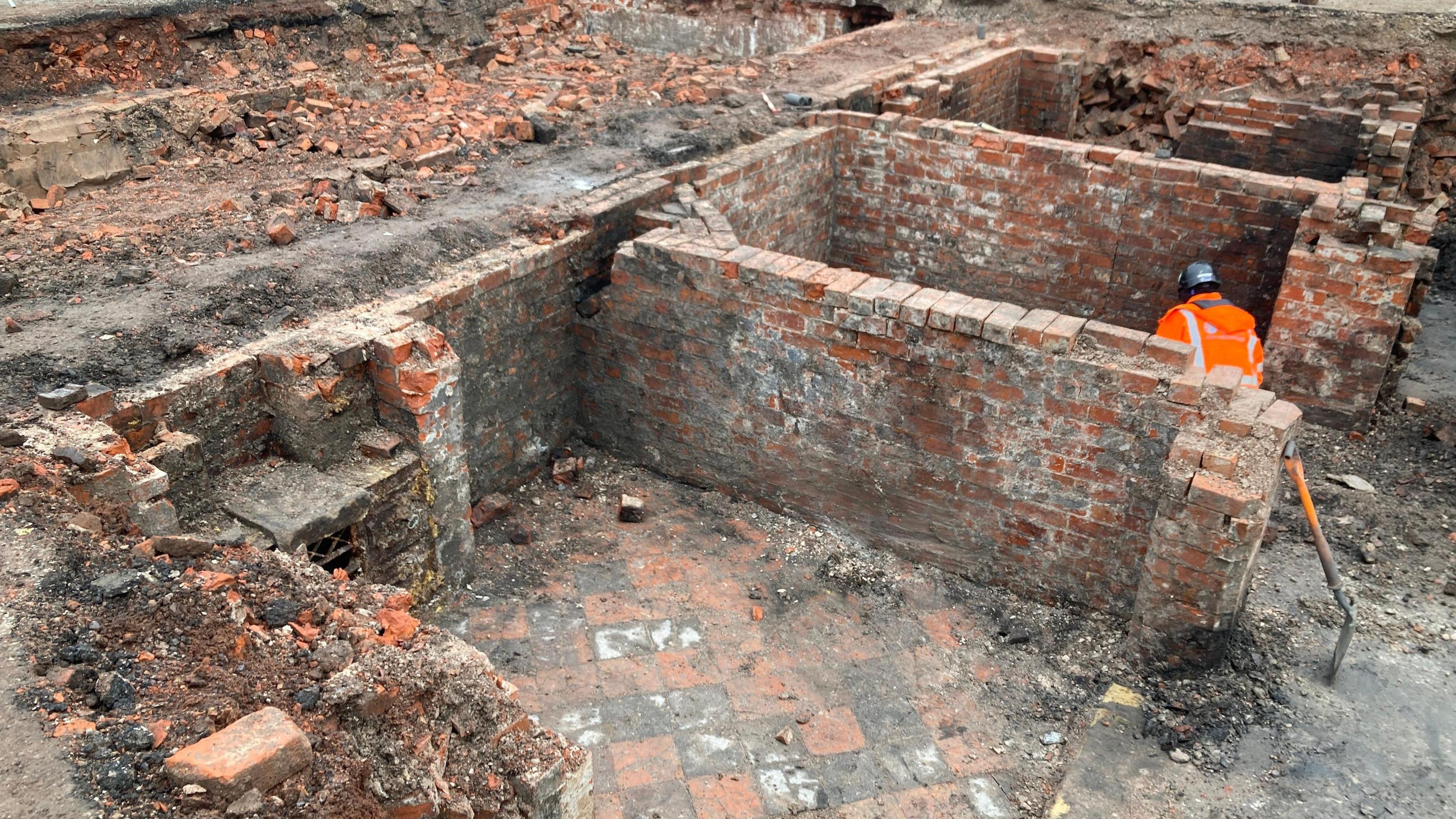 An excavation site where rows of brick walls are visible, a person in an orange hi-viz jacket and a helmet is visible behind one of the walls.