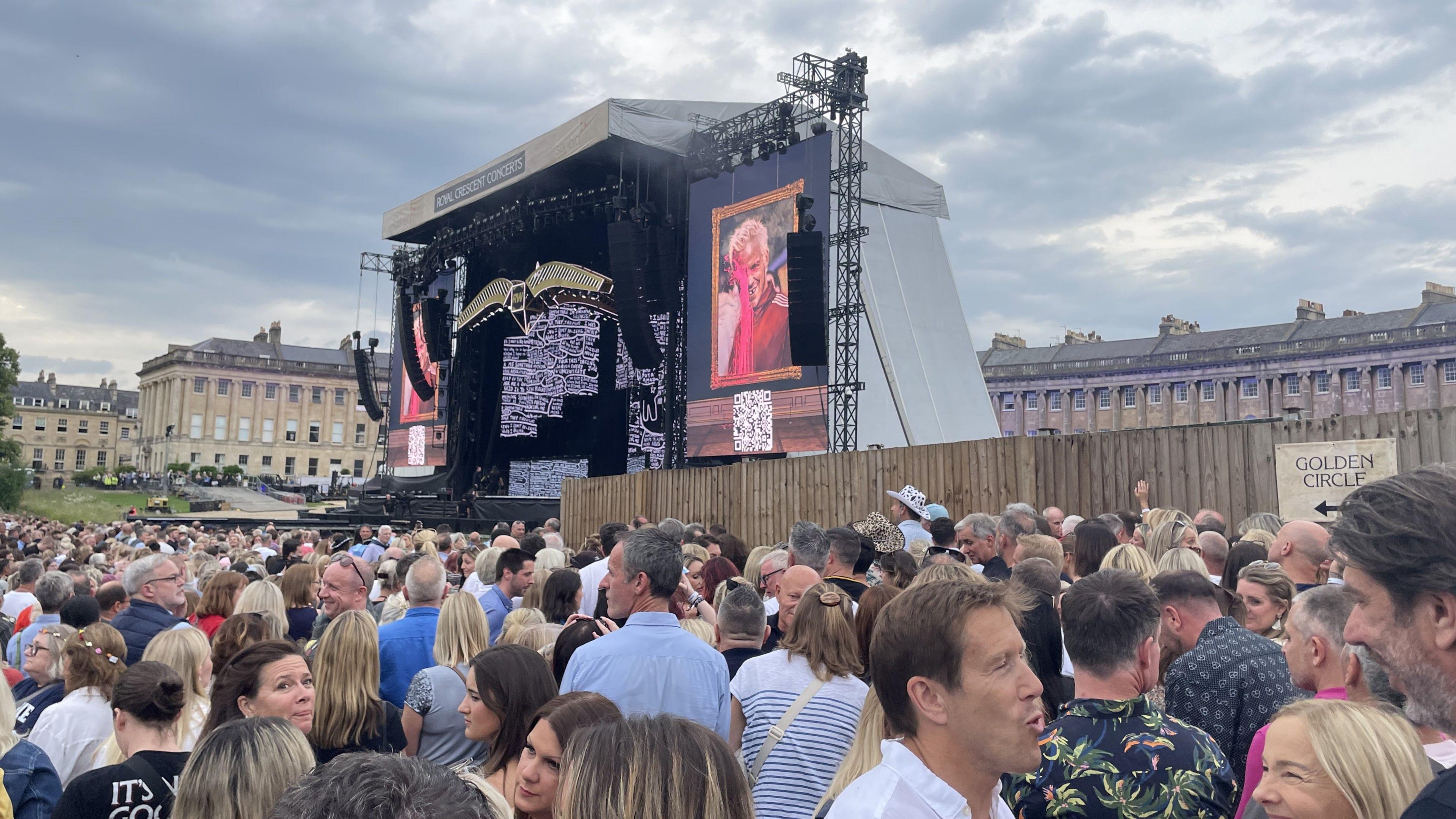 A large crowd of people standing near the front of the stage at the Royal Crescent in Bath. It is a cloudy day and everyone is stood around chatting as the music has not started yet.
