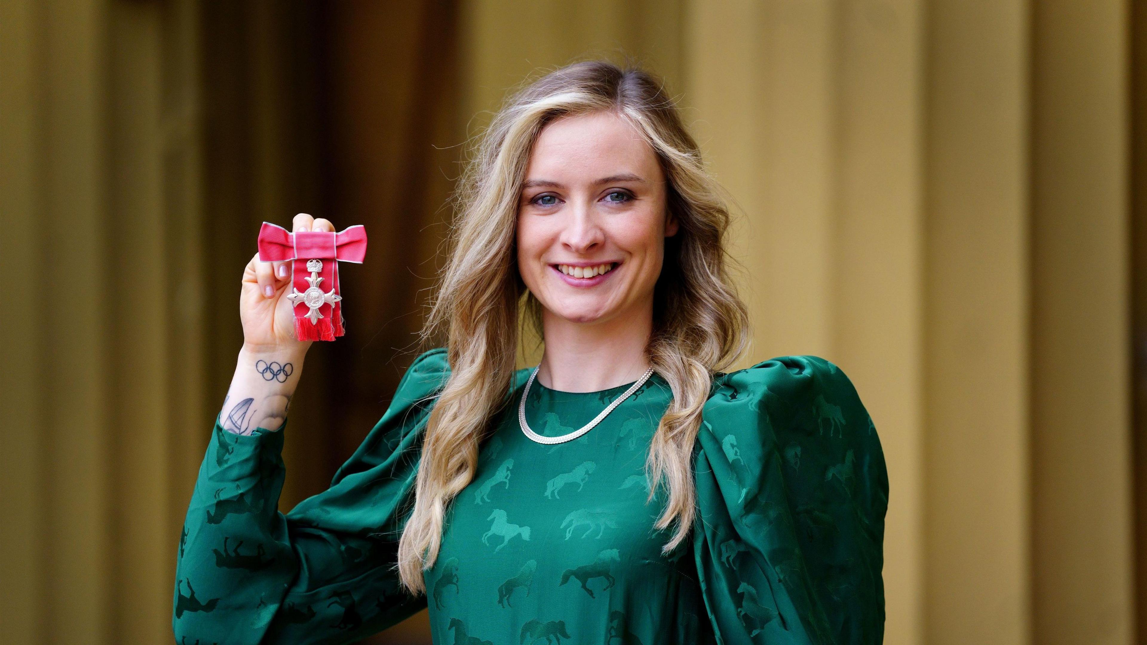 Charlotte Worthington with long brown hair, wearing a green dress and smiling whilst holding a medal in her right hand.  An Olympic tattoo is visible on her right hand. There are gold curtains behind her.