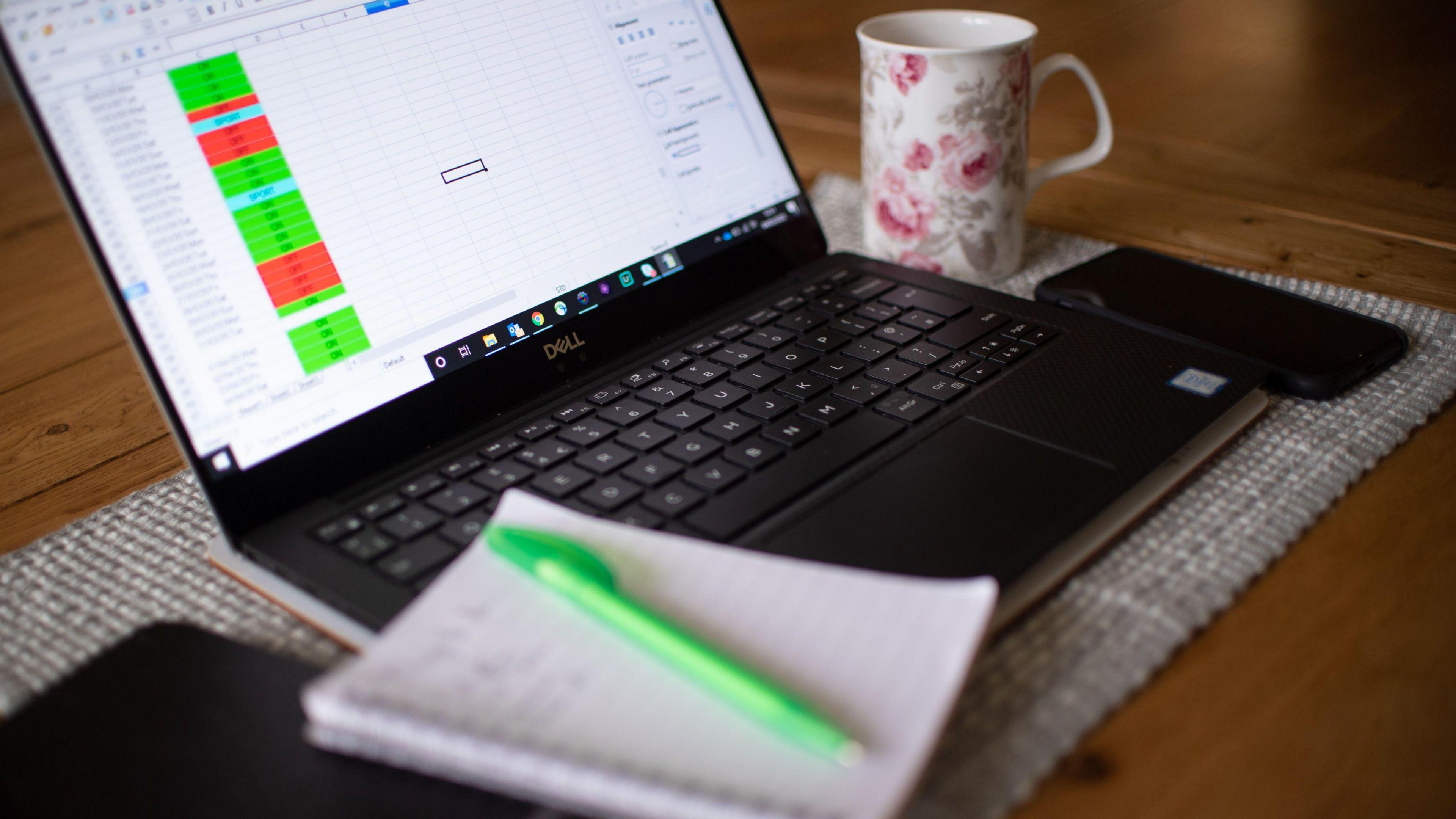 A laptop with a spreadsheet on it is open with a white ring-bound notepad resting on its keyboard. A coffee cup with a pink floral design on it and a black mobile phone are next to the laptop.