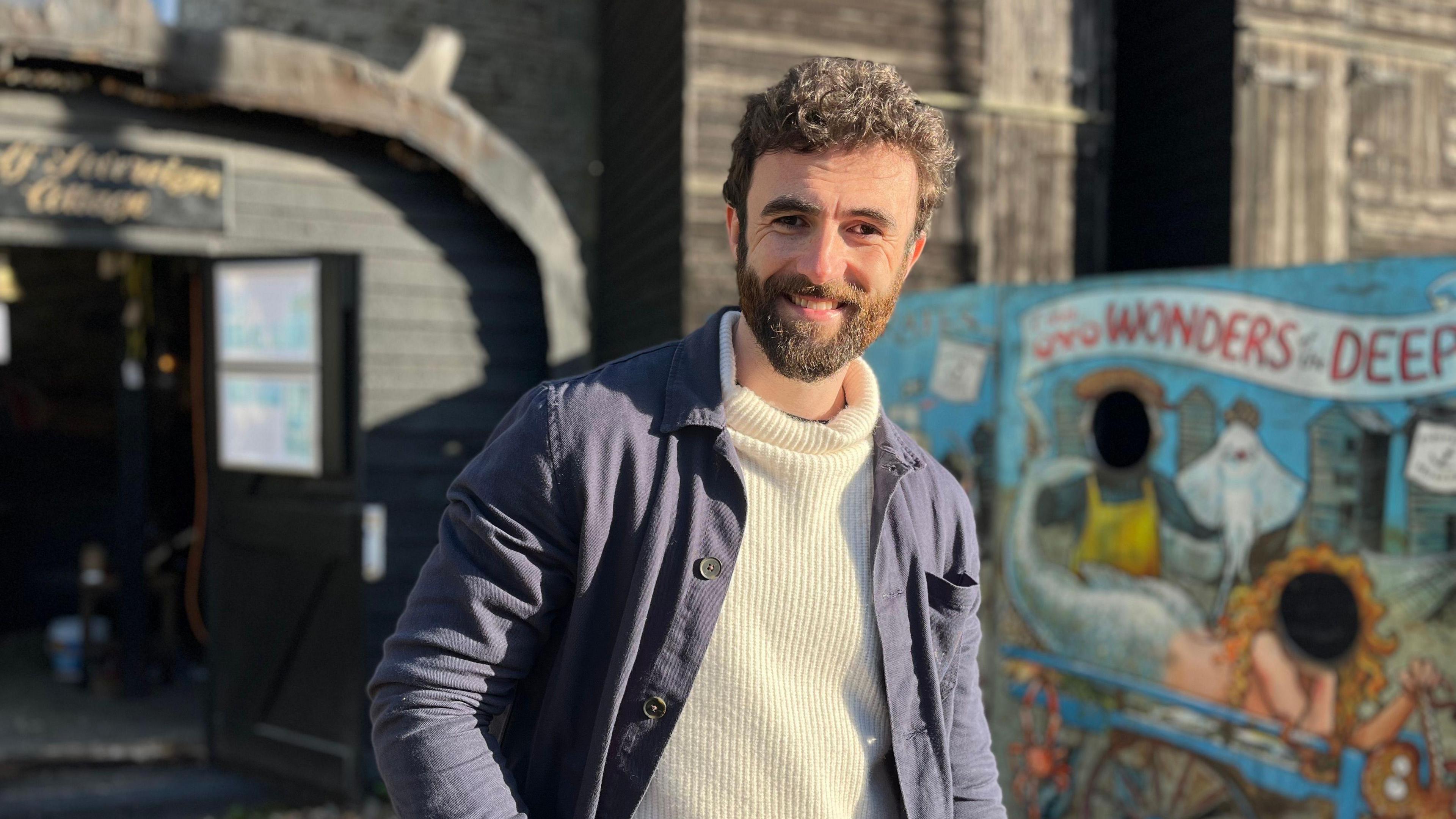 A man wearing a ribbed cream jumper and navy blue over shirt. He is stood in front of a fishermans hut and a seaside face cut-out stand and is smiling.