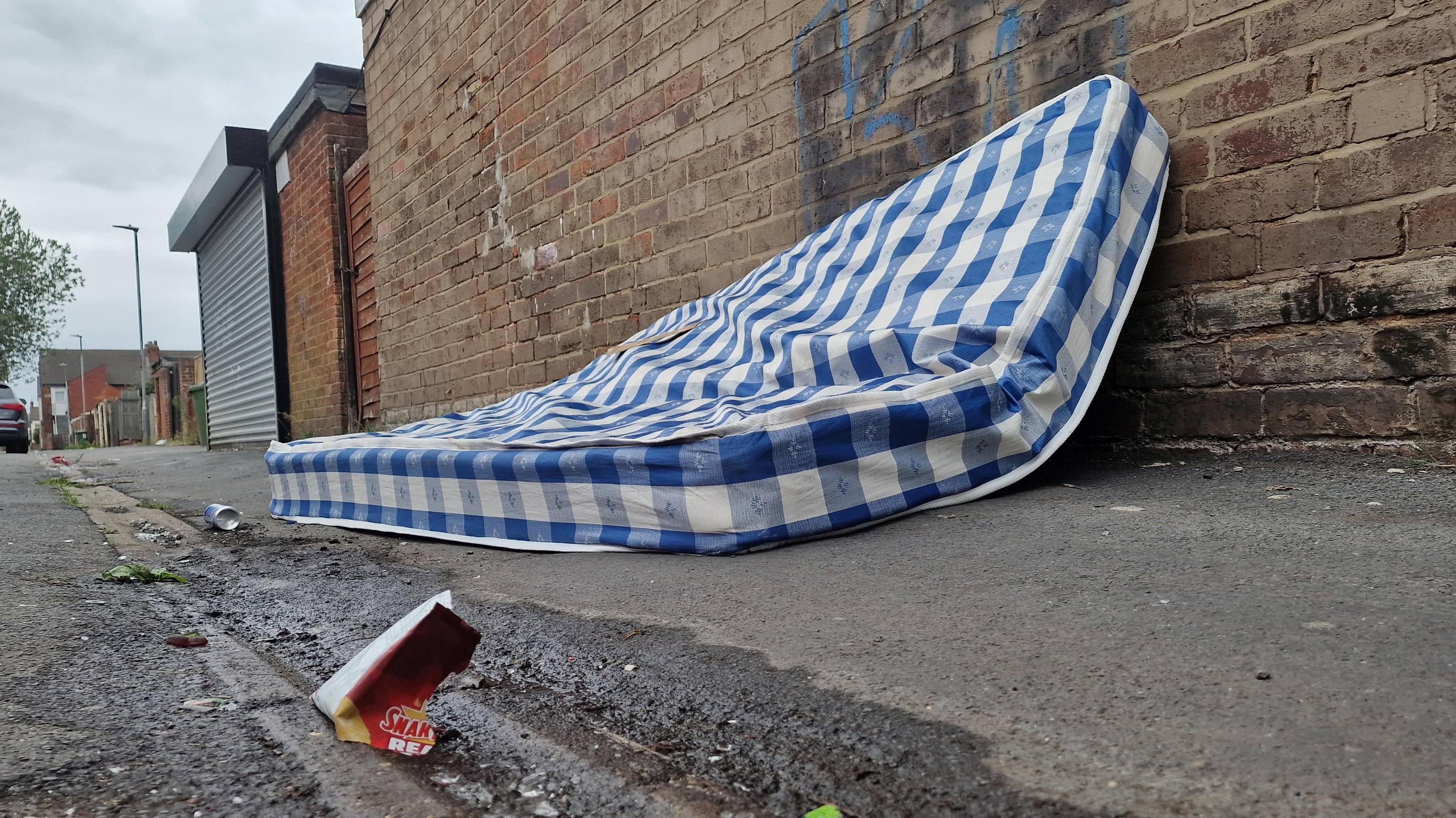 Blue and white mattress and some litter in an alleyway in front of a brick building.