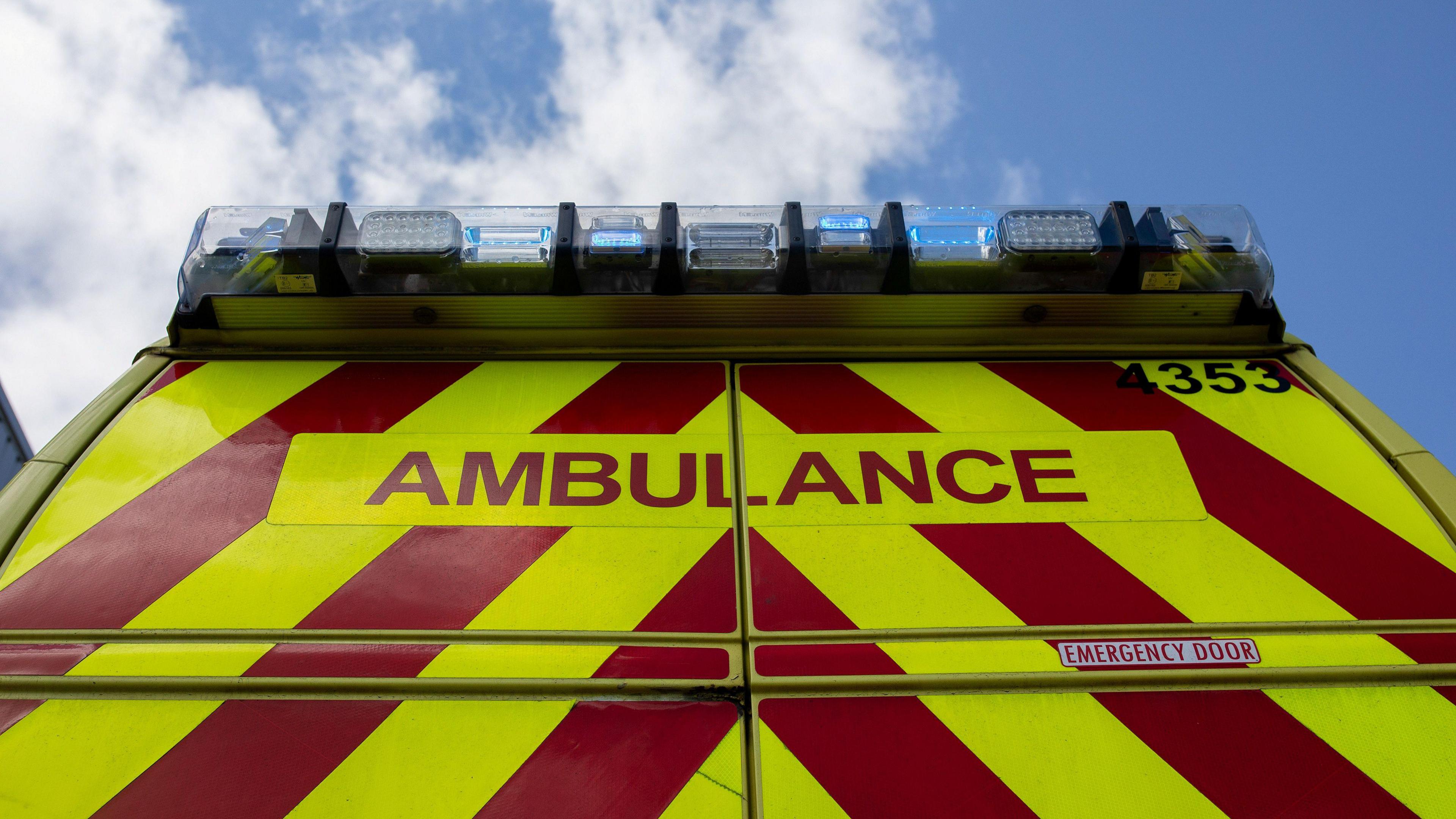 The back of an ambulance showing the yellow and red chevrons and the word "ambulance" in red letters. On top of the vehicle are blue lights, which are switched on. In the background, blue sky and white clouds are visible.