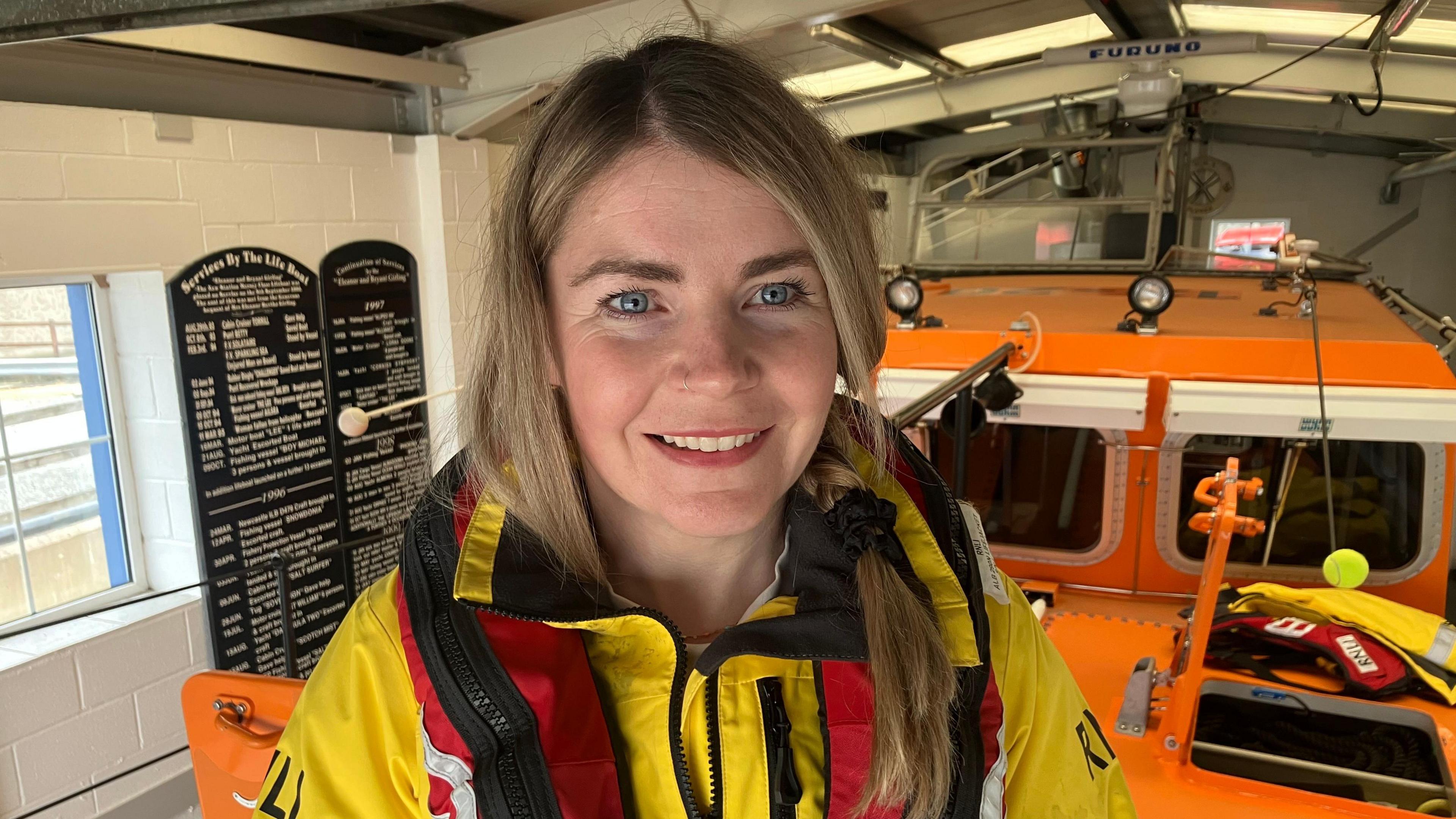 Caoimhe is standing in front of an orange boat with a yellow lifeboat crew jacket and red life jacket on. She has blond long hair in a ponytail and blue eyes. She is smiling at the camera.