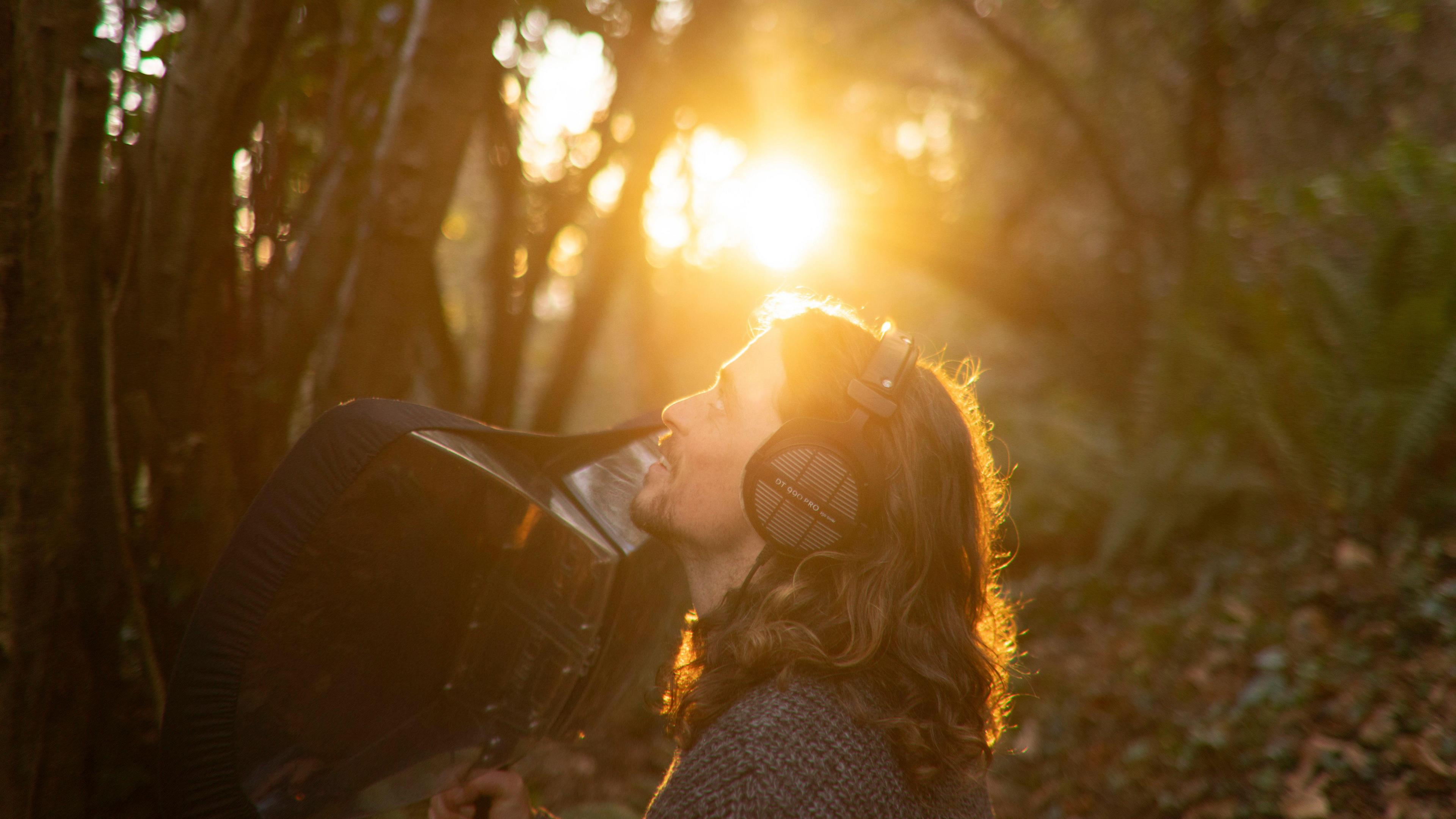 A long-brown haired man looks to the sky. He is surrounded by shrubs and holds a large black sound recorder, that resembles an umbrella. He wears black headphones, a black t-shirt and a blue jumper.