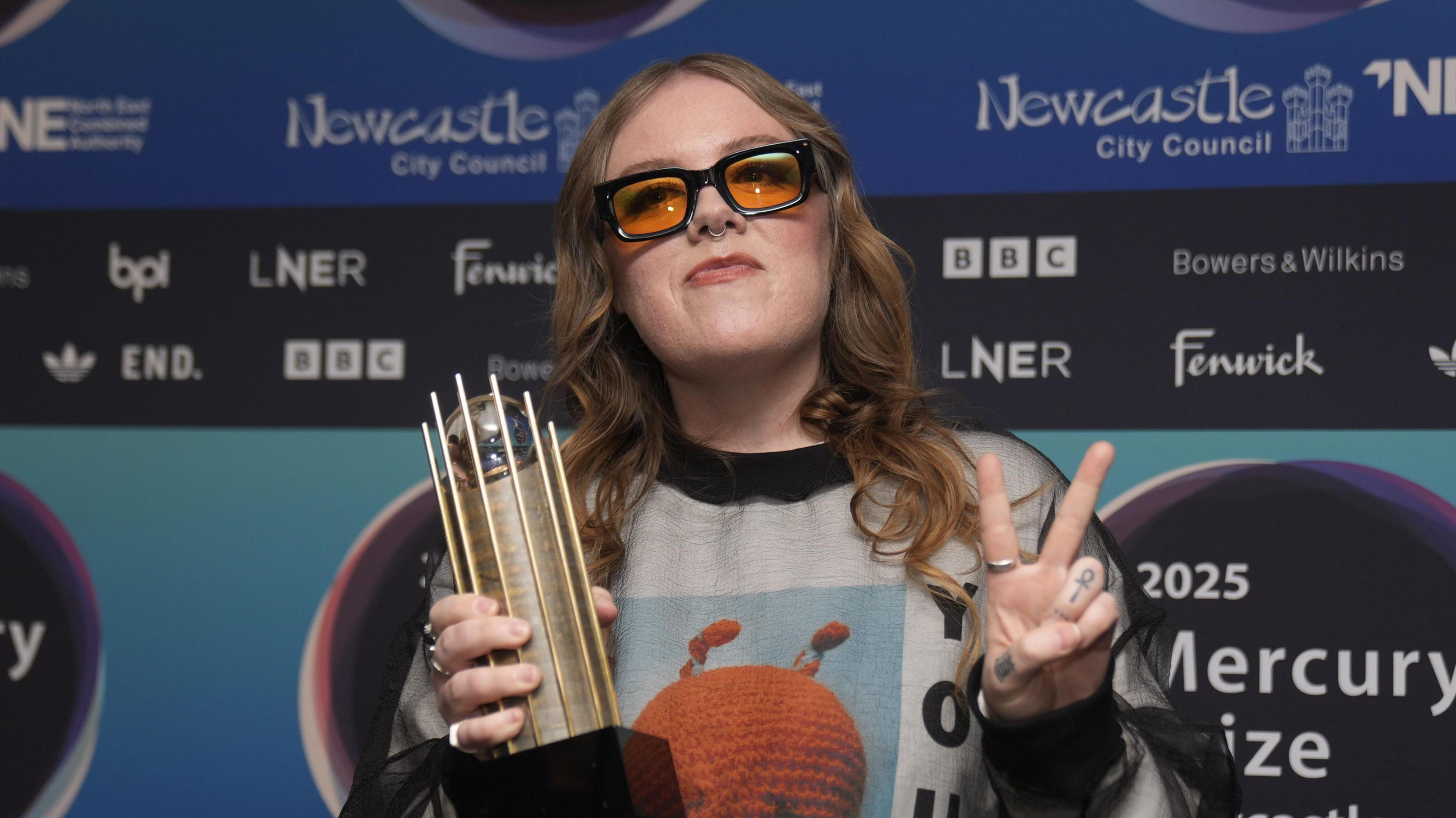 Emma-Jean Thackray posing with her Mercury nomination trophy outside the Utilita Arena. She has long mousey-blonde hair and is wearing orange-tinted glasses and a nose ring. She is holding her fingers up in a peace sign.