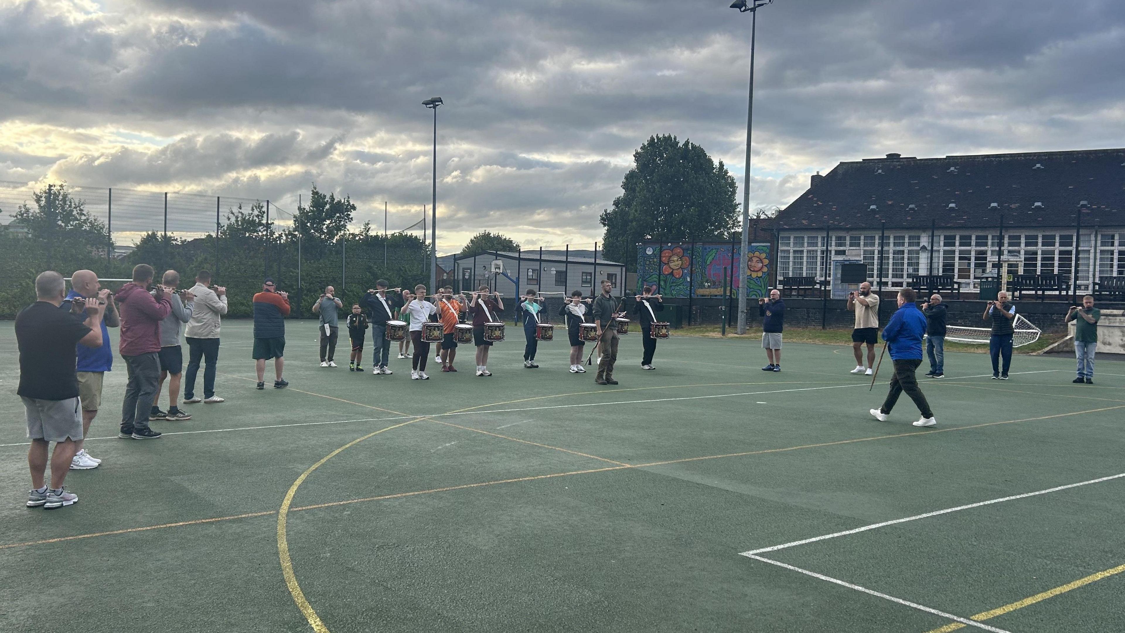 There are around 20 people shown rehearsing their band music. There are those playing the flute and drummers also. The picture is a wide shot of the band members standing on a football pitch.