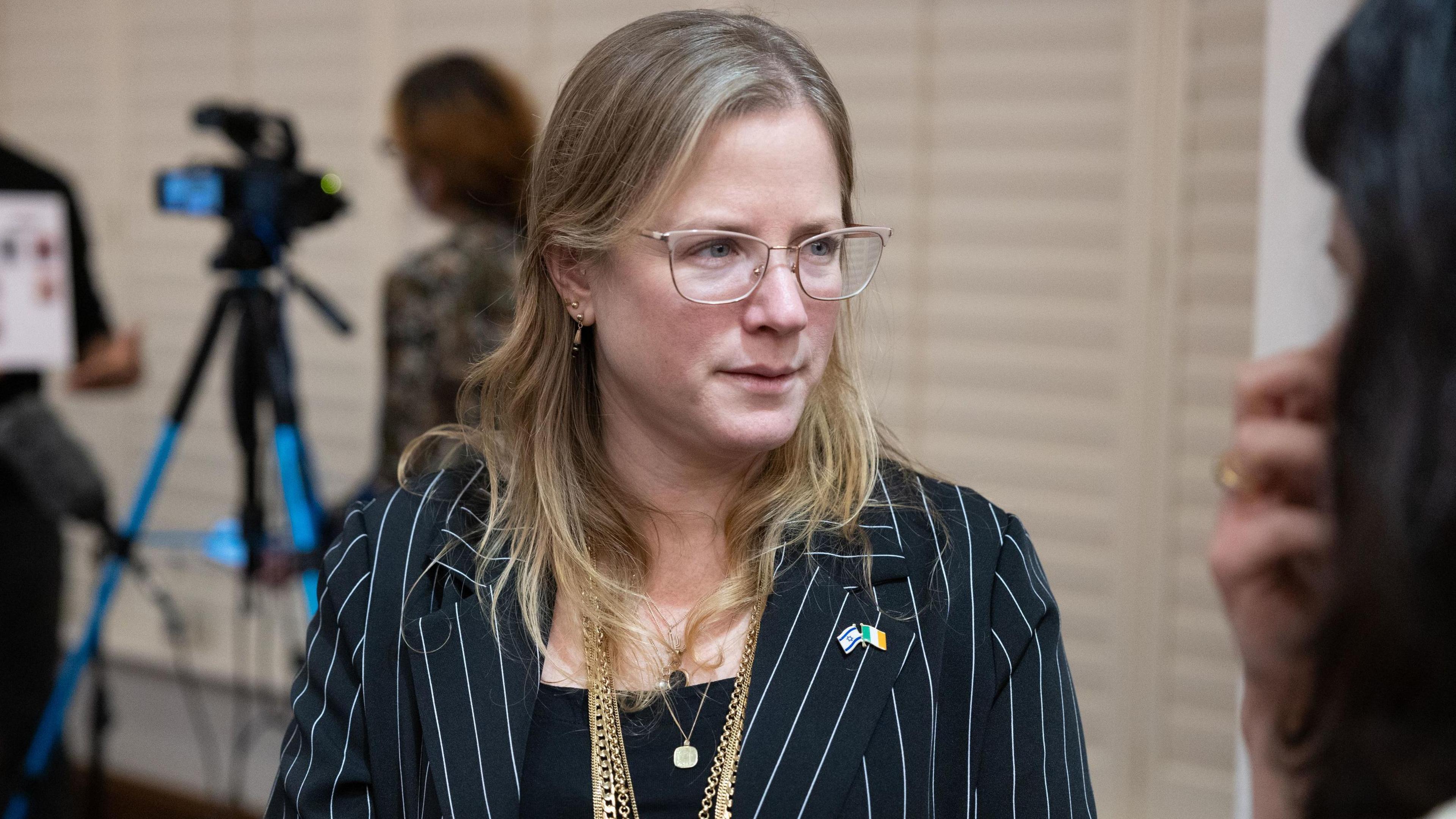 Dana Erlich wears a black suit with skinny white pinstripes. She has on four gold chains around her neck and a black top underneath her suit. She has blonde hair and glasses. On her lapel is an Israel flag and Ireland flag. Behind her, out of focus, is a brown haired women with a black camera. 