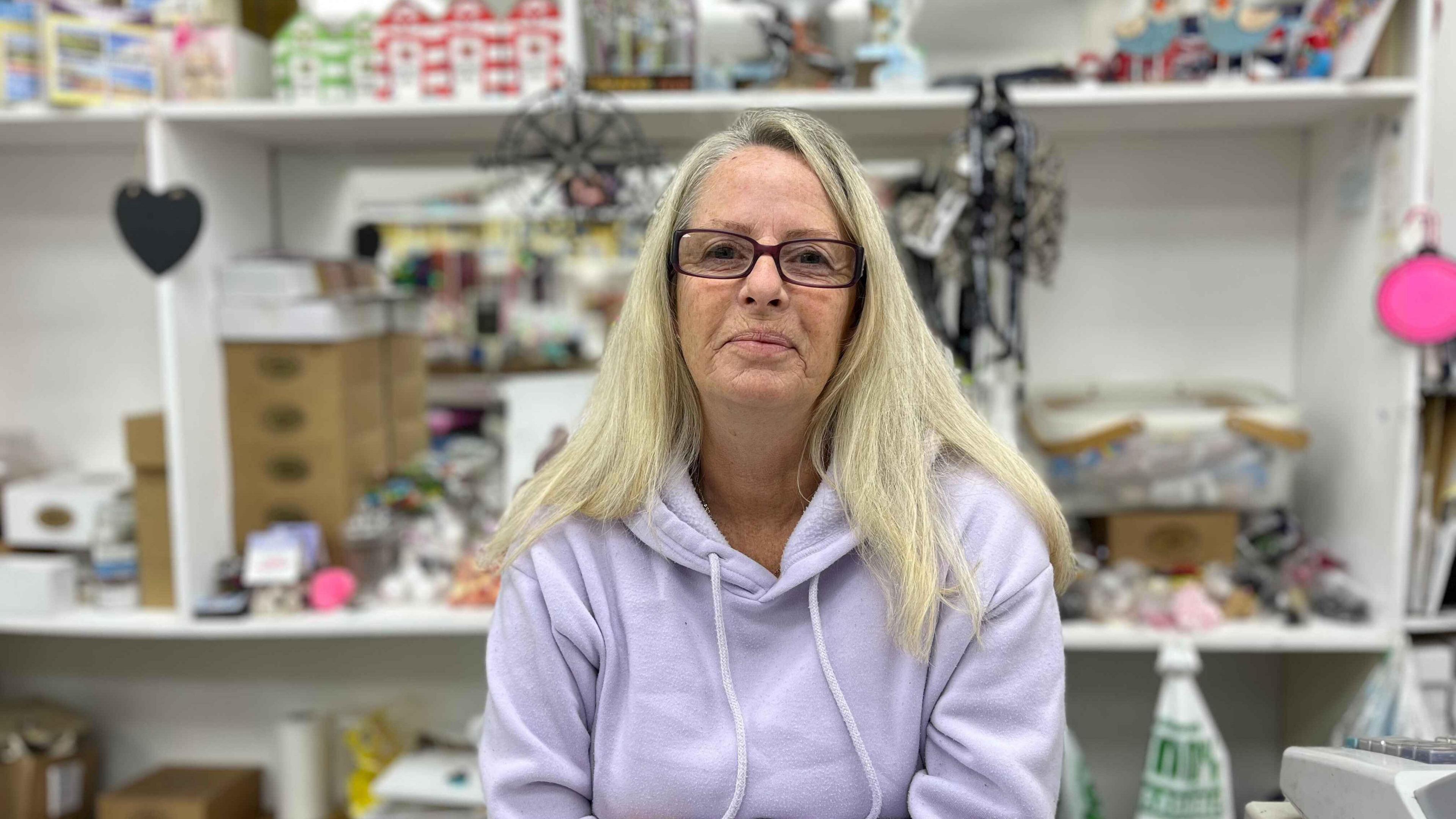 Heidi Durrant wearing a light purple jumper and has blonde hair. She is standing behind the counter with toys, sweets and other stock behind her