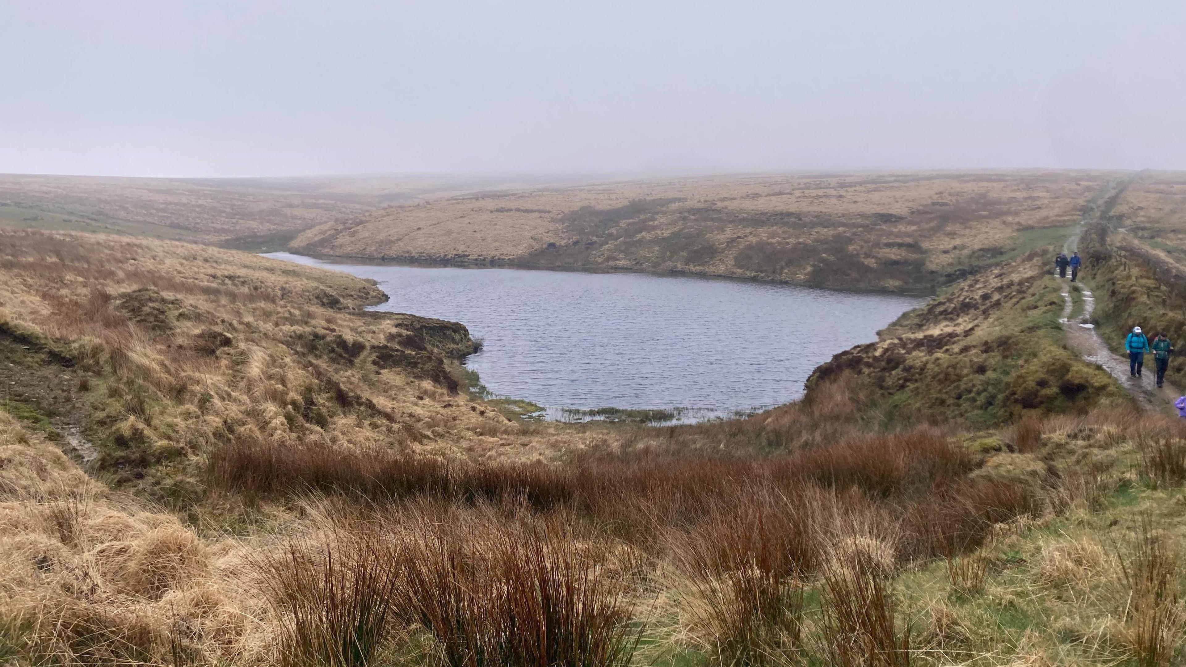 People walk along a lakeside path near The Chains area of Exmoor on a wet and cloudy day. The clouds are grey and lying low. The lake is surrounded by moorland which looks dry.