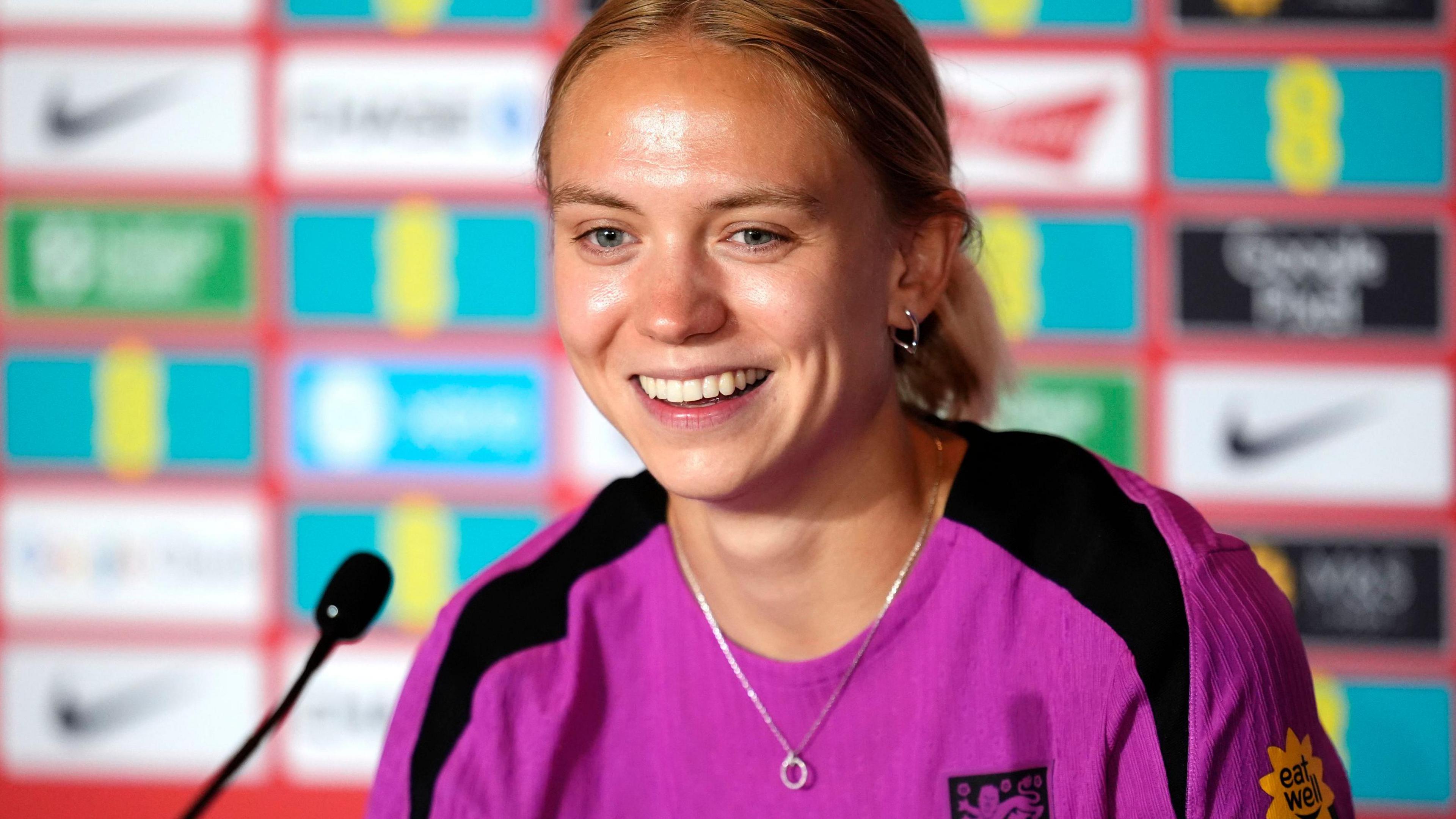 Esme Morgan smiles as she sits in front of a microphone wearing a pink England training top. She has blonde hair tied back in a pony tail, a silver hoop earing in her left ear and a silver necklace. She is sat in front of a blurry background featuring various sponsorship logos.
