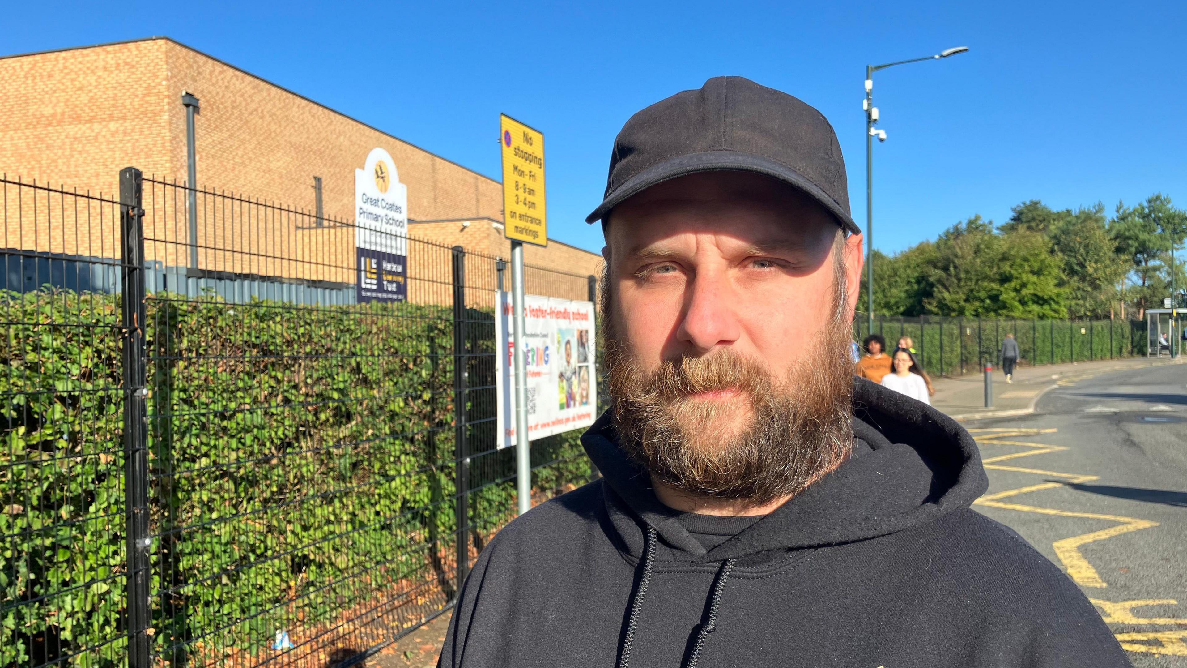 A man with a bushy beard wears a plain black cap and black hoodie. He is standing in front of a modern brick school building, which is surrounded by a black wire fence and green hedge. A yellow parking sign reads: "No stopping Mon - Fri, 8-9am, 3-4 pm on entrance markings". Yellow zig-zag lines can be seen on the road. Another sign advertises fostering services. More people can be seen in the background.